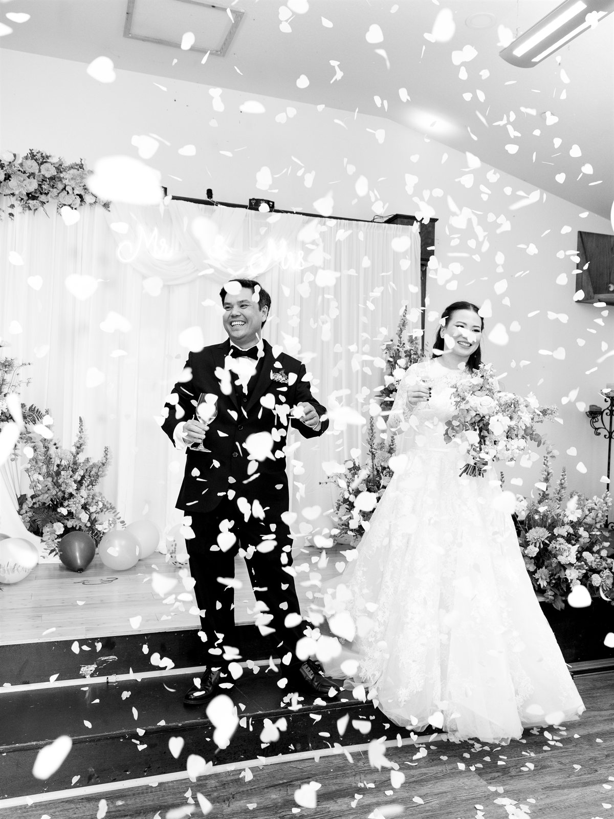 Bride and groom toasting through a shower of heart confetti at their reception, black and white