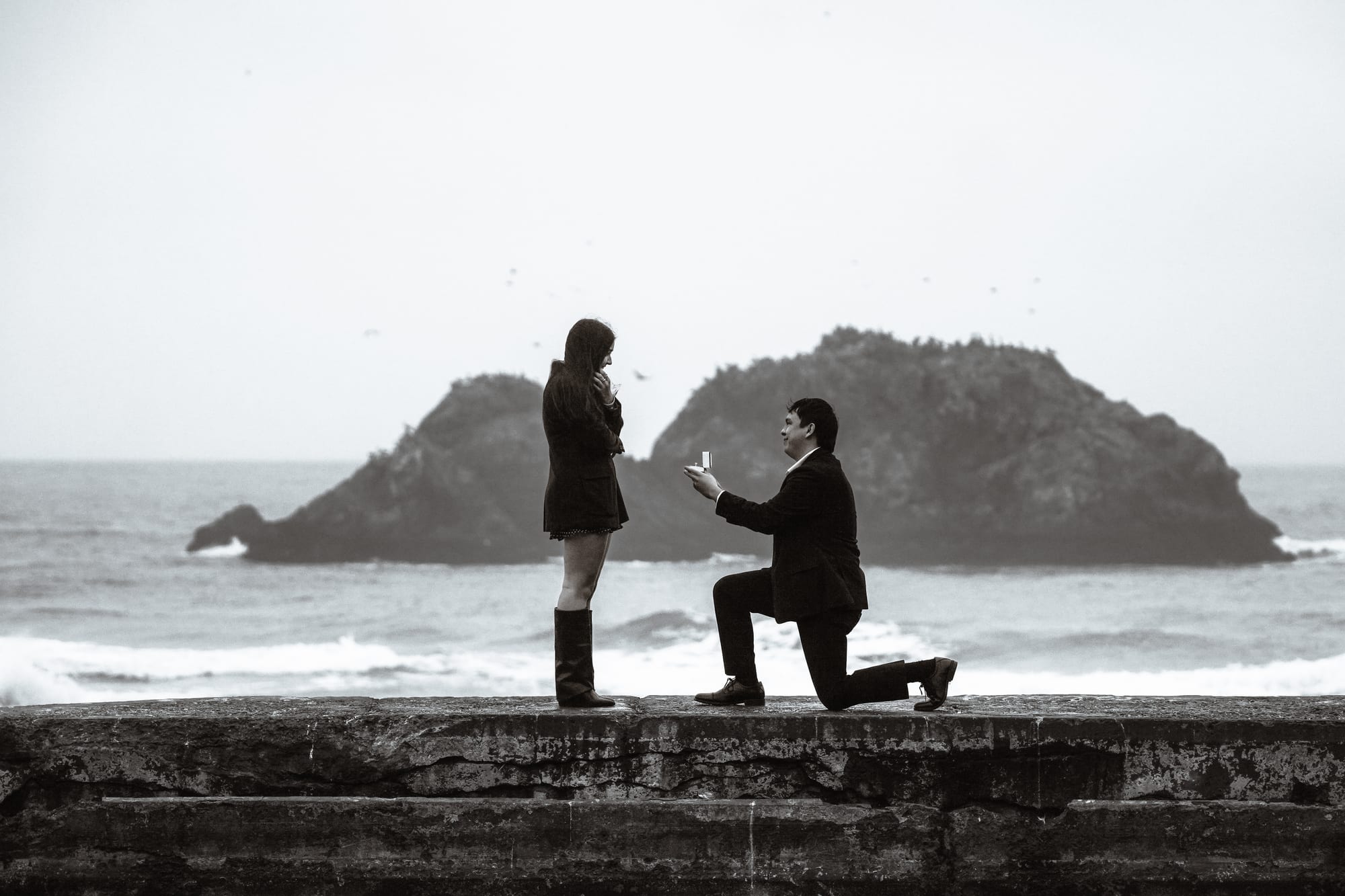 Black-and-white shot of him on one knee holding out the ring box as she covers her mouth in surprise, the twin sea stacks behind them