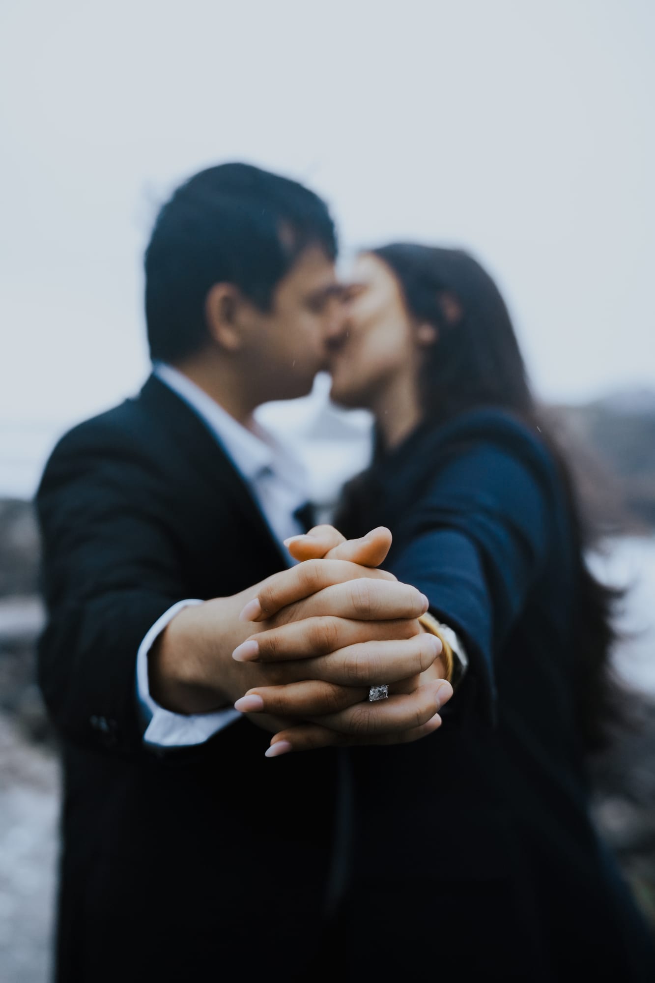 Close-up of their clasped hands as they kiss, her new ring catching the light between his fingers