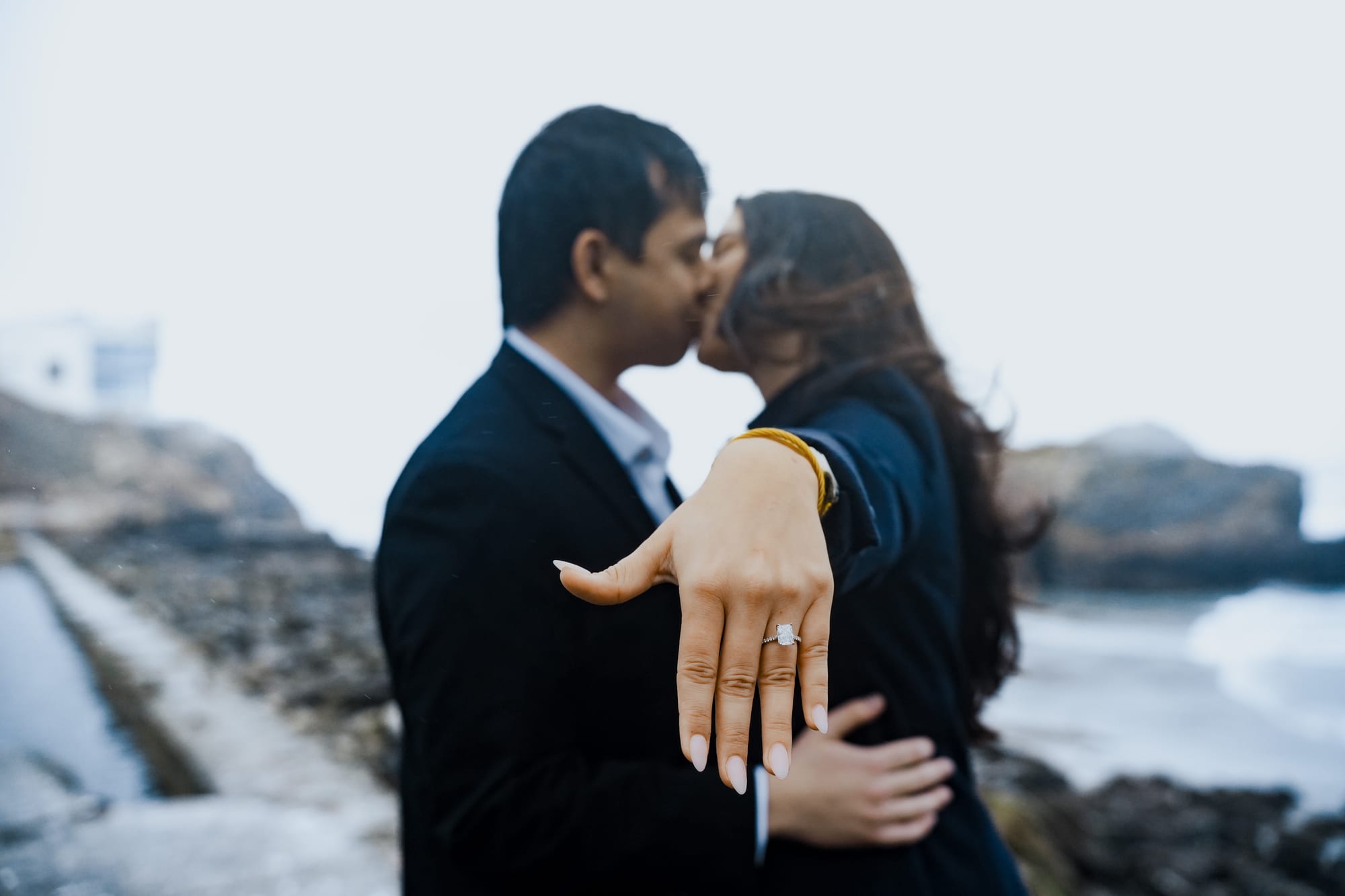They kiss as she holds her ring hand out to the camera, her hair blown across her face, the bluff-top building soft behind them