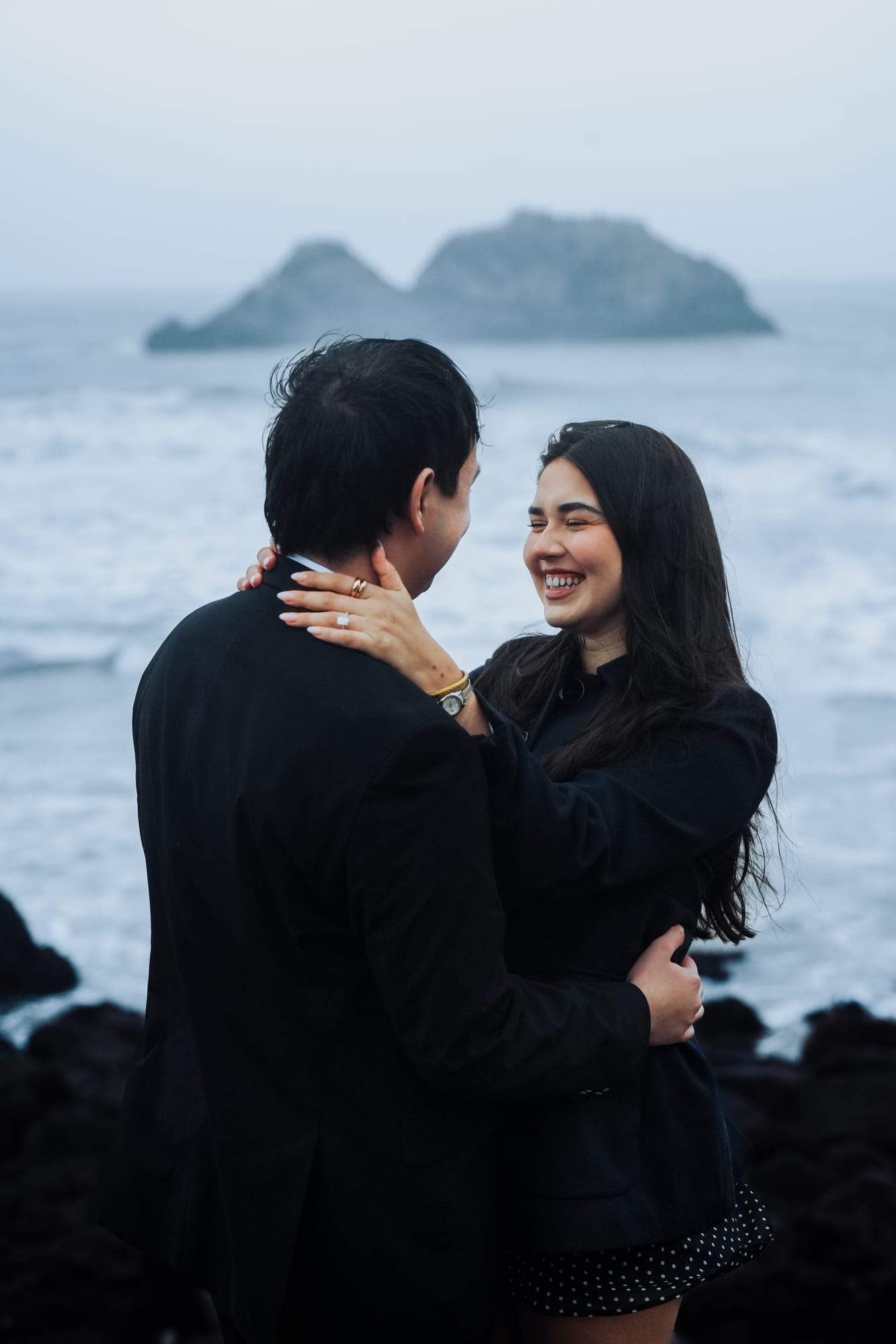 She laughs with her hand on his shoulder, her new ring and gold bracelet on display, the ocean and sea stacks behind them