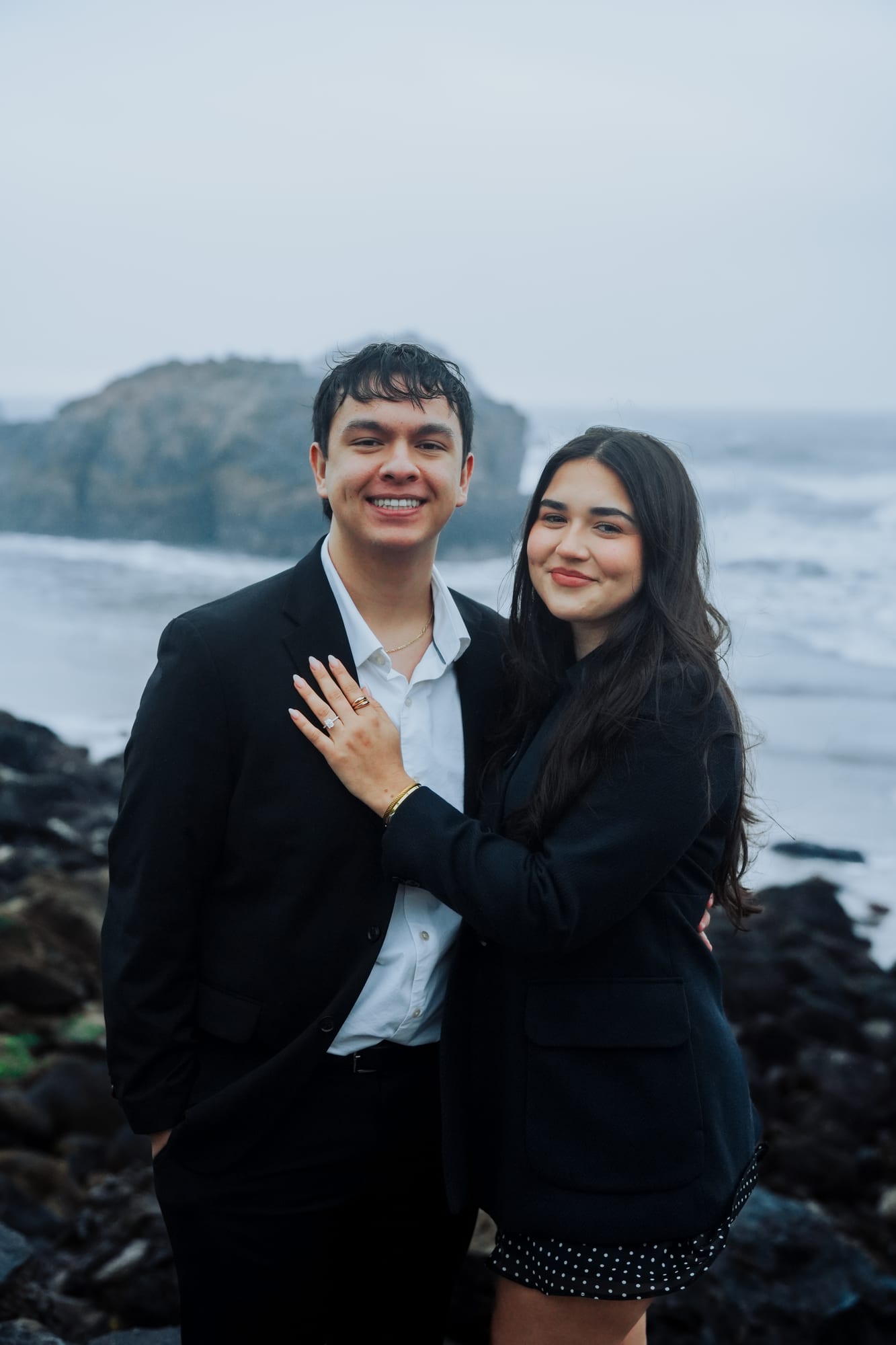 Portrait of the newly engaged couple smiling at the camera, her ring hand resting on his chest, the rocky coastline behind them
