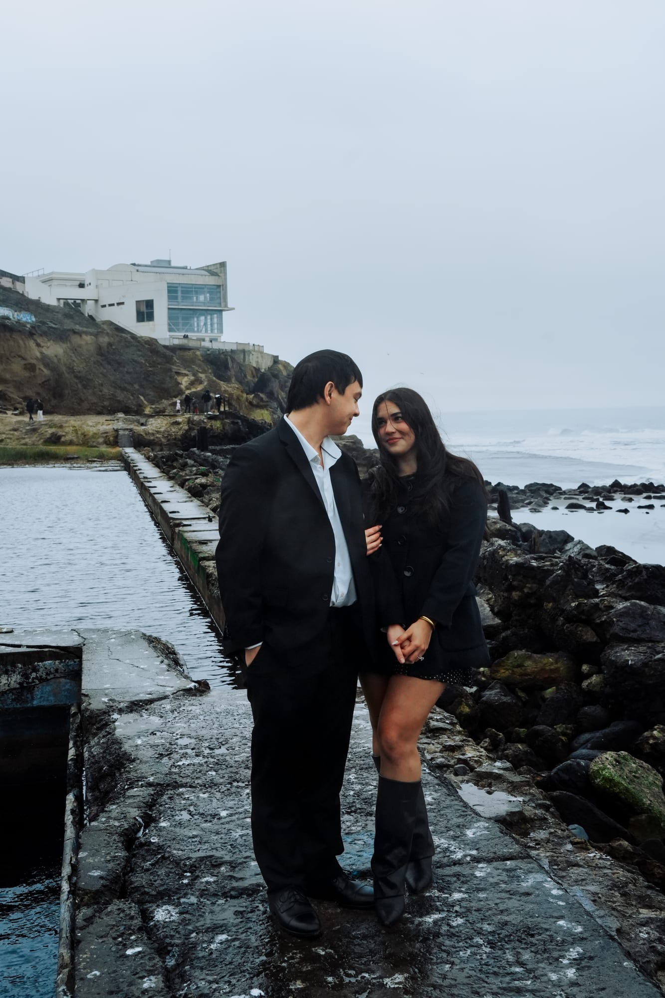 Couple standing close together at the edge of the old bath ruins, the cliffside building perched on the bluff behind them