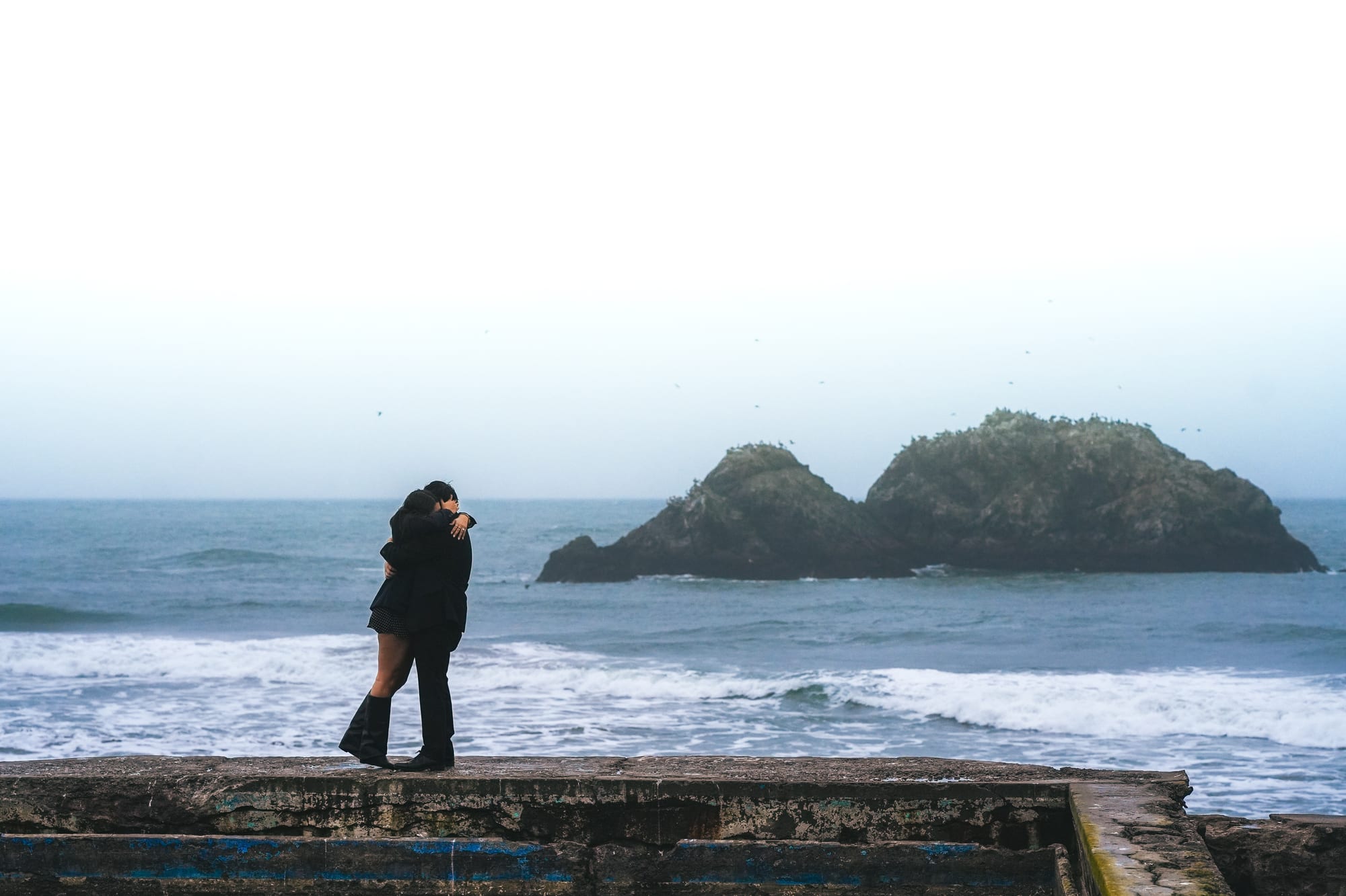 Wide shot of the couple embracing on the seawall, the twin sea stacks and crashing surf filling the frame behind them