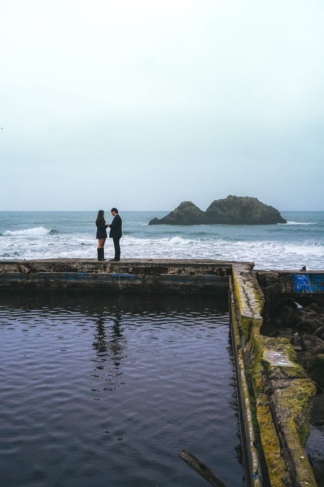 Wide shot of the couple standing hand in hand at the edge of the ruins, their reflection in the still water below, sea stacks in the distance