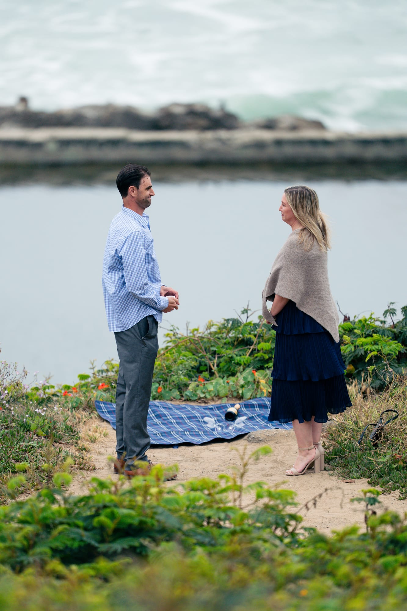 Man and woman standing face to face on a coastal bluff, a plaid picnic blanket and champagne laid out at their feet, waves breaking below