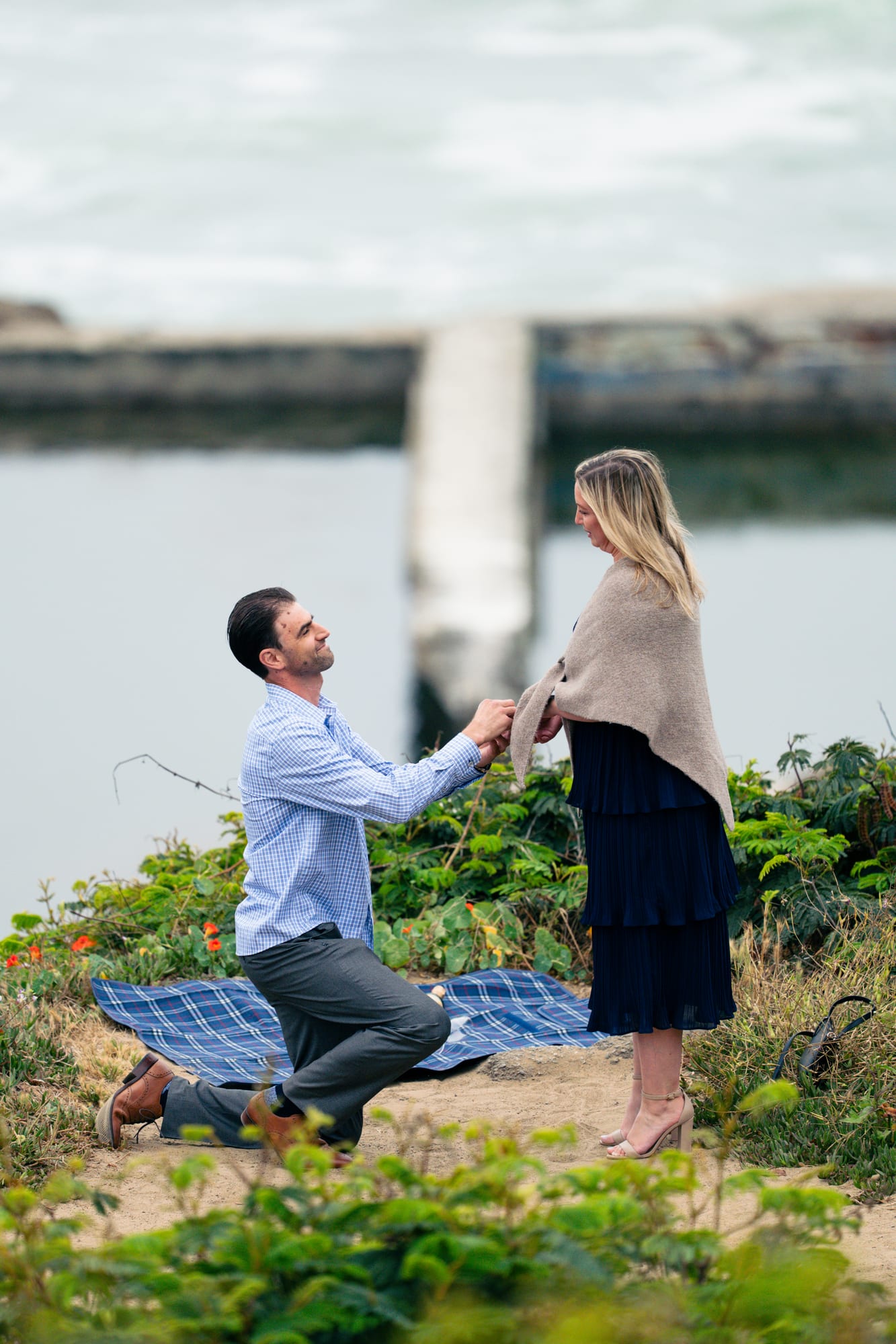 Man kneeling on one knee on the picnic blanket, holding the woman's hand mid-proposal above the shoreline