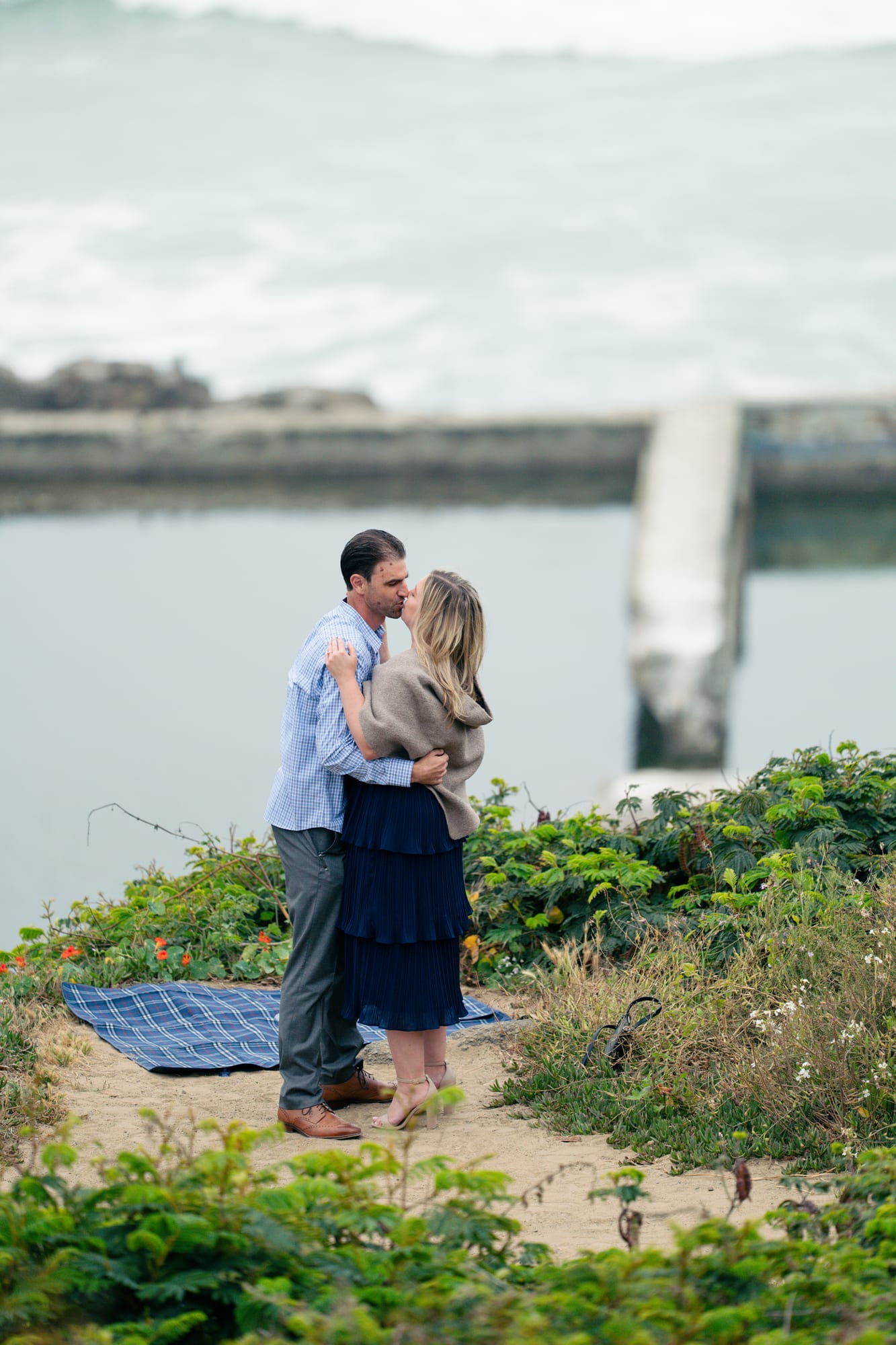 The newly engaged couple kissing on the bluff, her taupe wrap around her shoulders, the tide rolling in behind them