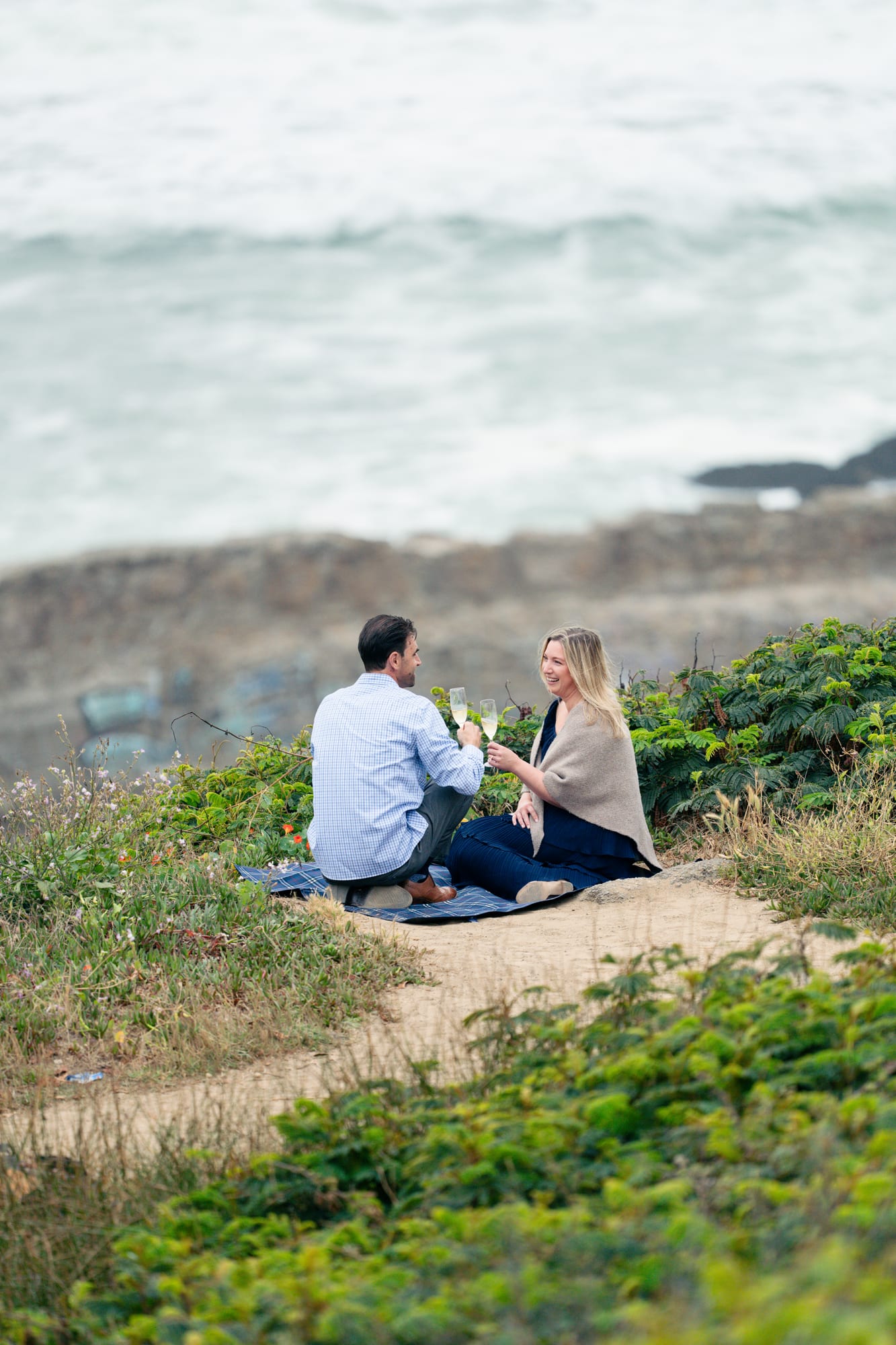 Couple seated on the picnic blanket toasting with champagne flutes, she's laughing, the rocky shoreline spread out behind them