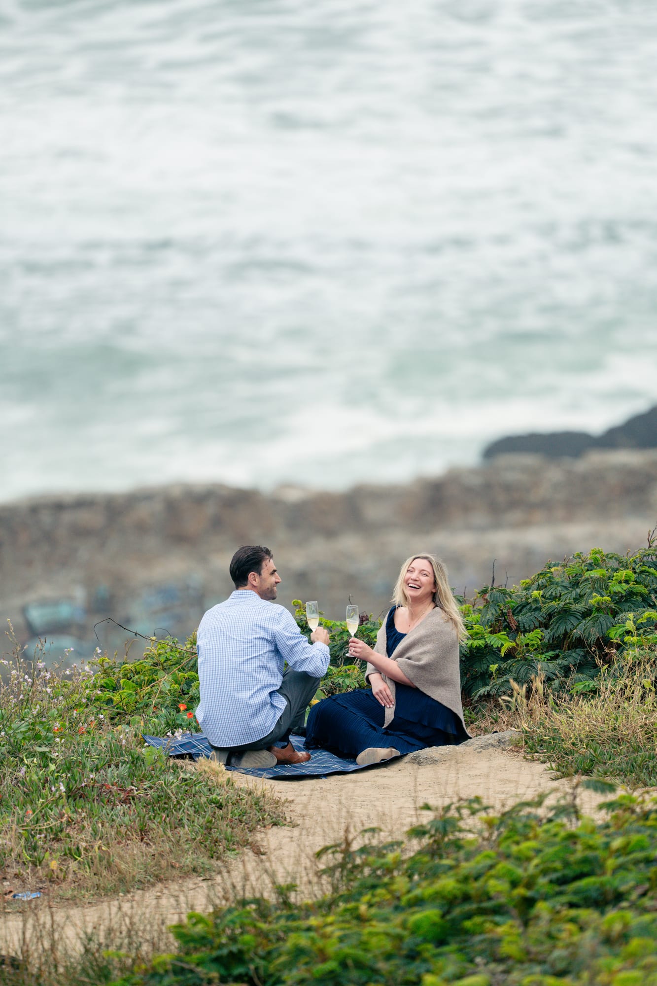 Couple seated back to camera on the blanket, raising champagne glasses in a toast over the fog-covered coast