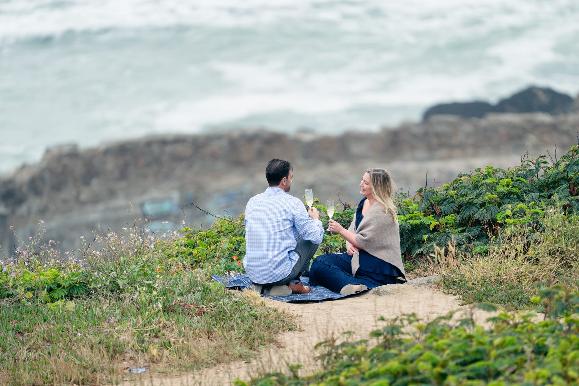 Wide view of the couple seated cross-legged on the picnic blanket, glasses raised, dwarfed by the cliffs and open water