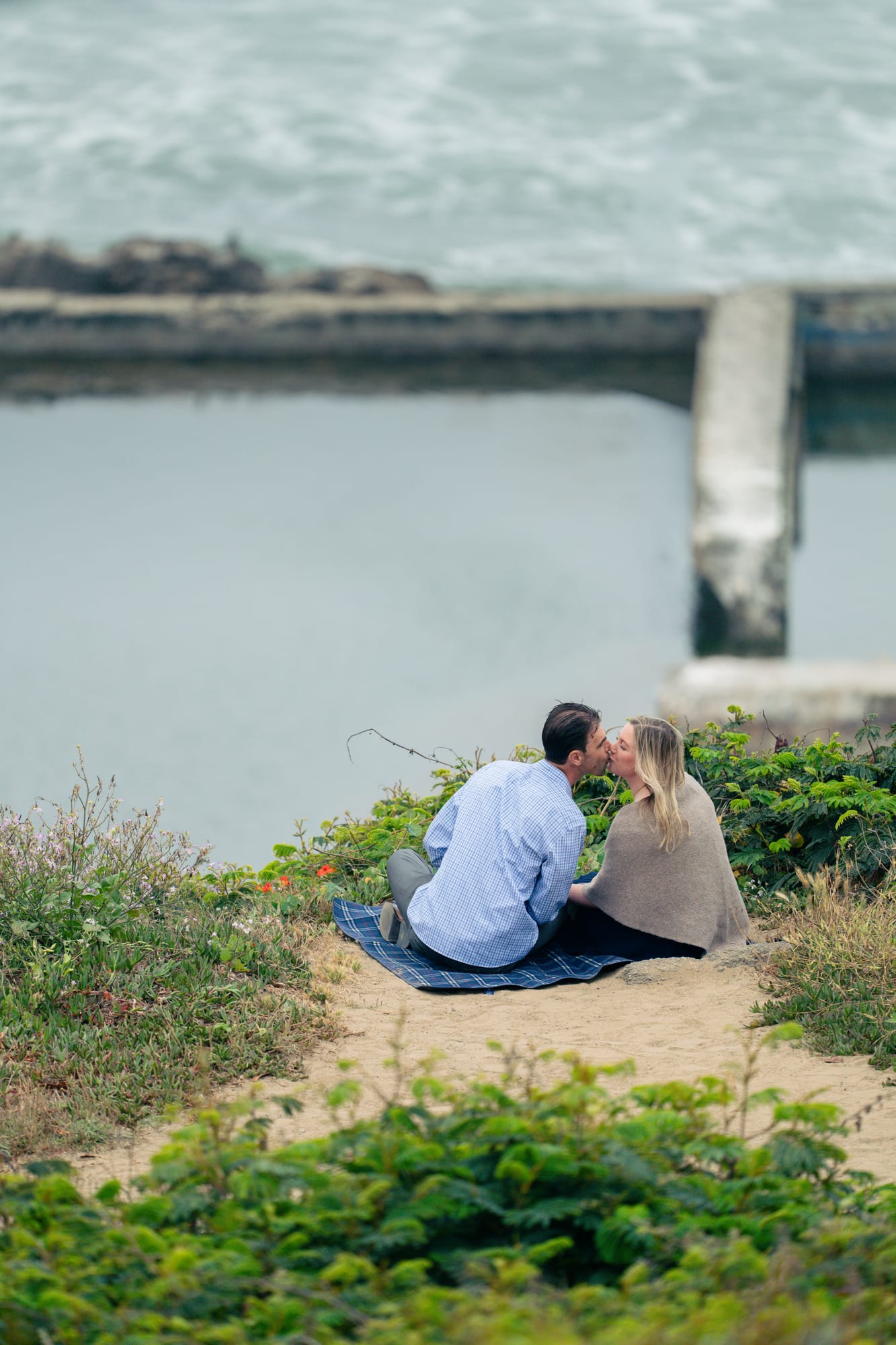 Couple kissing seated on the blanket, backs to camera, a concrete piling rising from the water behind them
