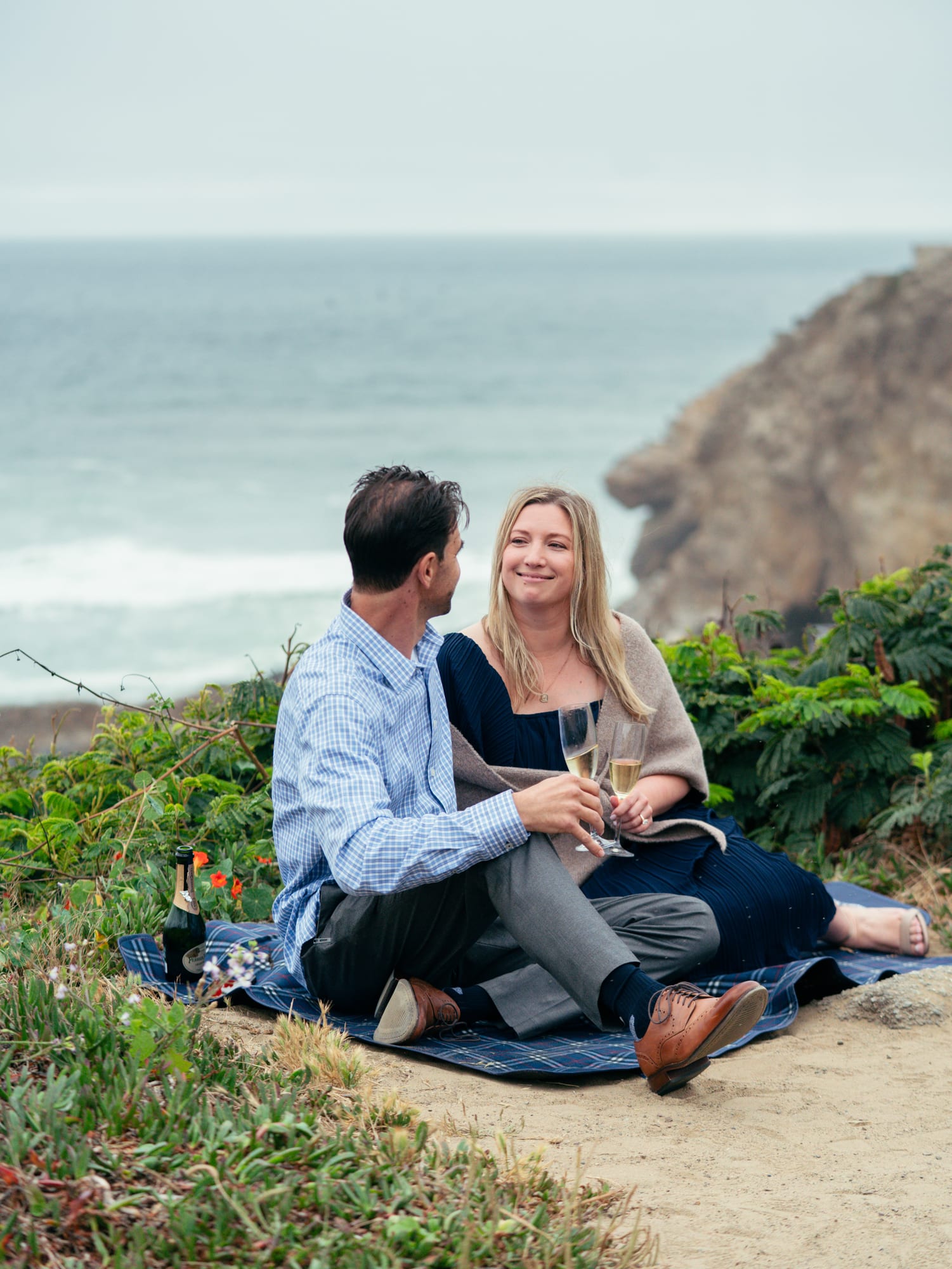 Couple seated close on the blanket, champagne bottle and wildflowers beside them, sea cliffs rising in the distance
