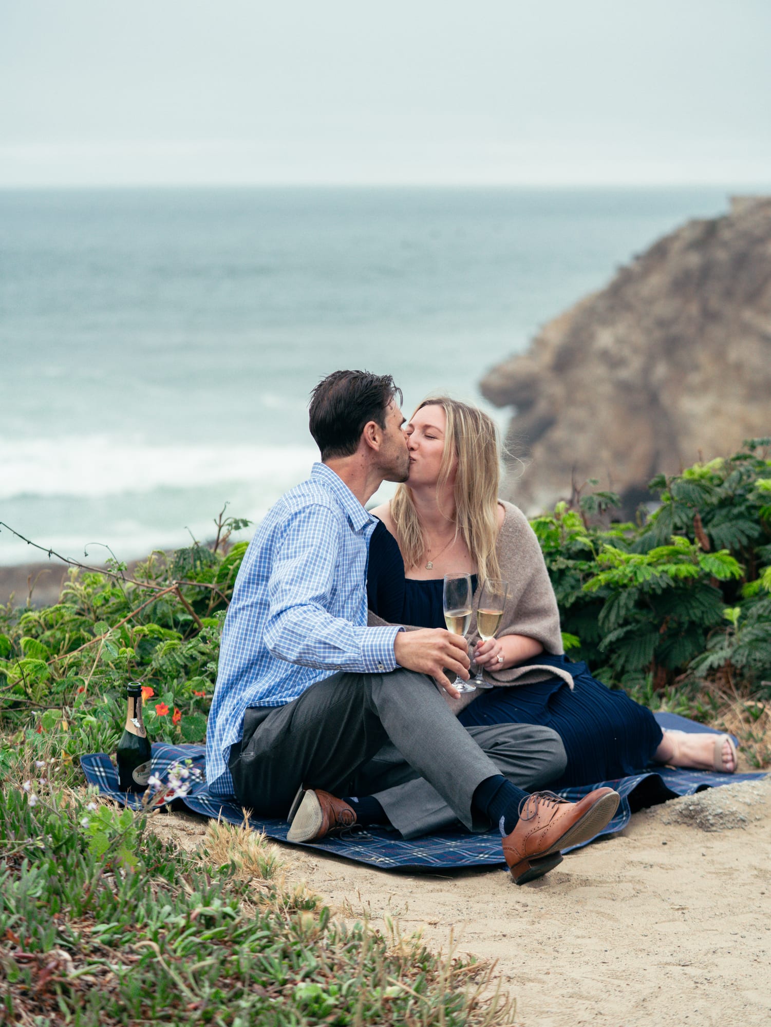 Couple kissing on the blanket, the champagne bottle resting in the grass beside them, a sea stack framed over her shoulder