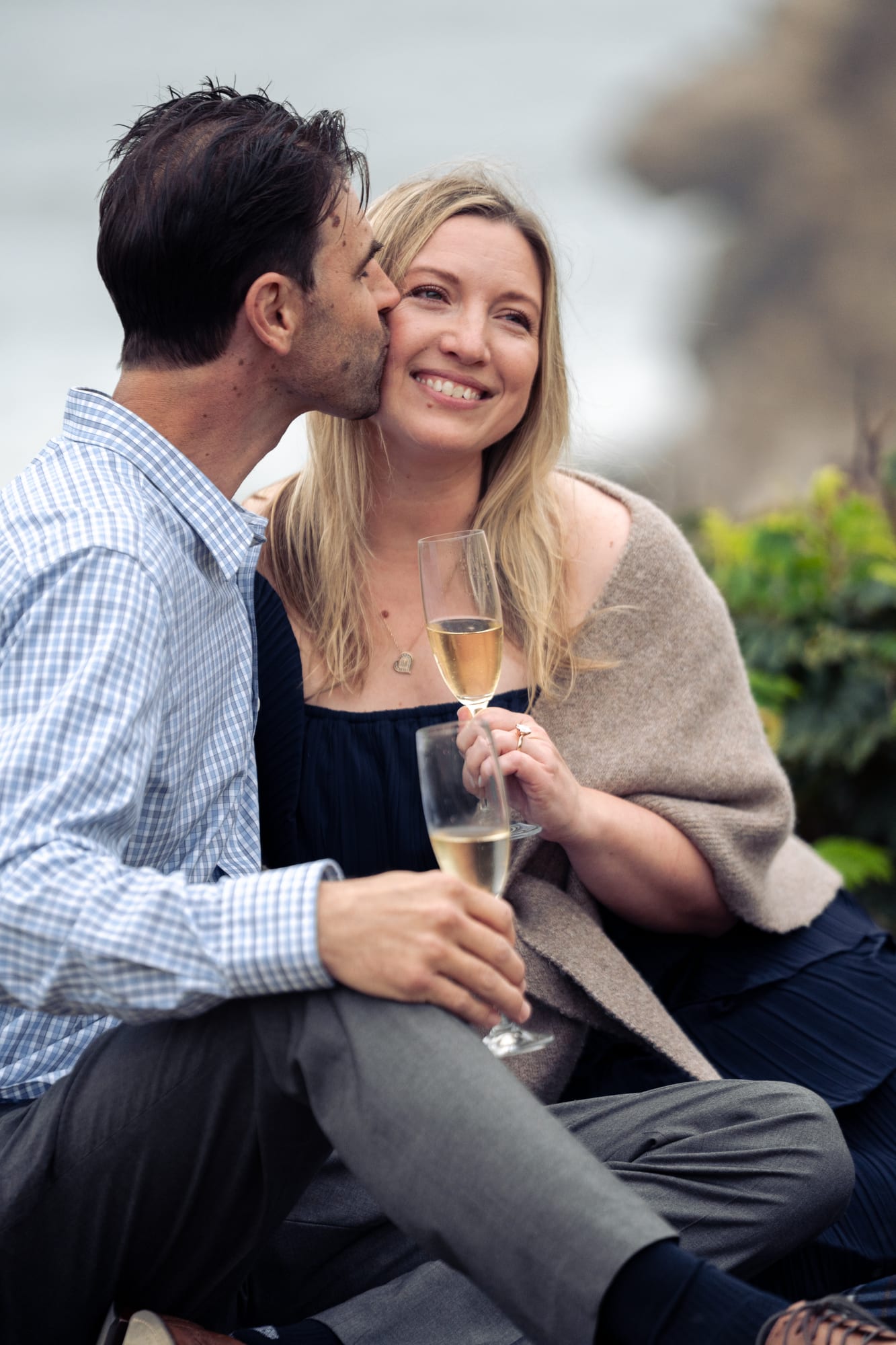 He kisses her cheek as she laughs, her new ring bright against the stem of her champagne flute