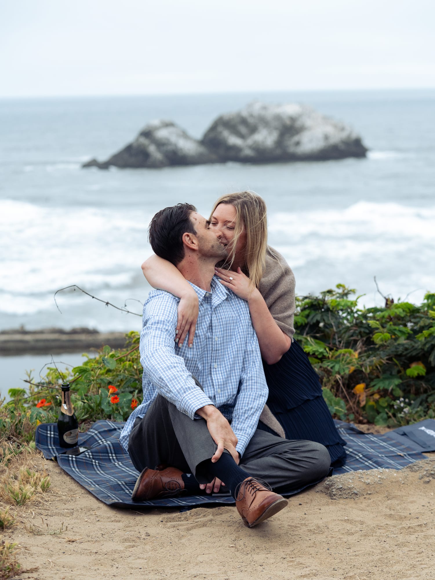 Couple laughing together on the blanket, her arms around his neck, the tide pools spread out below