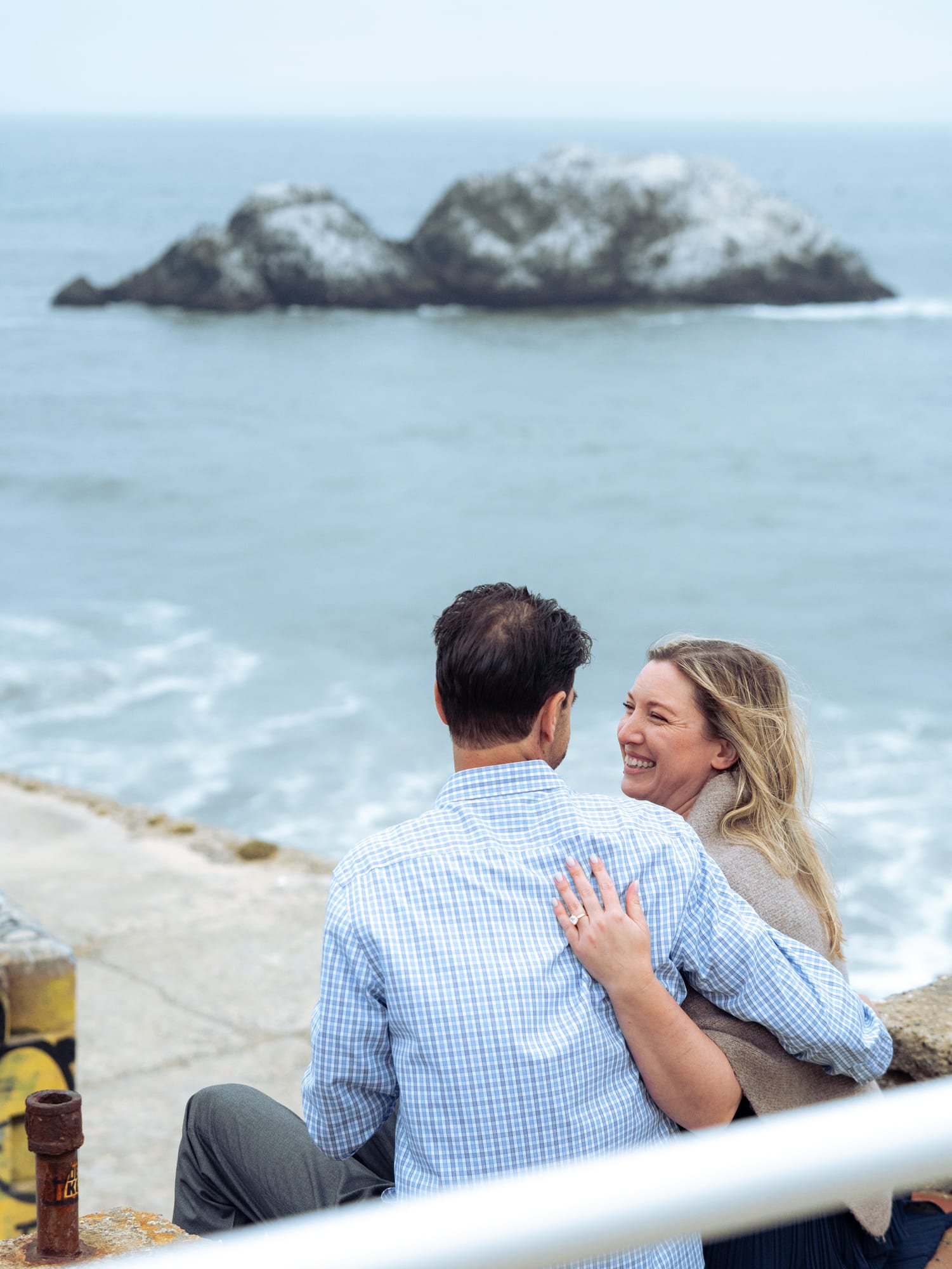 Couple embracing from behind on the concrete overlook, her ring hand on his shoulder, sea stacks out past the point