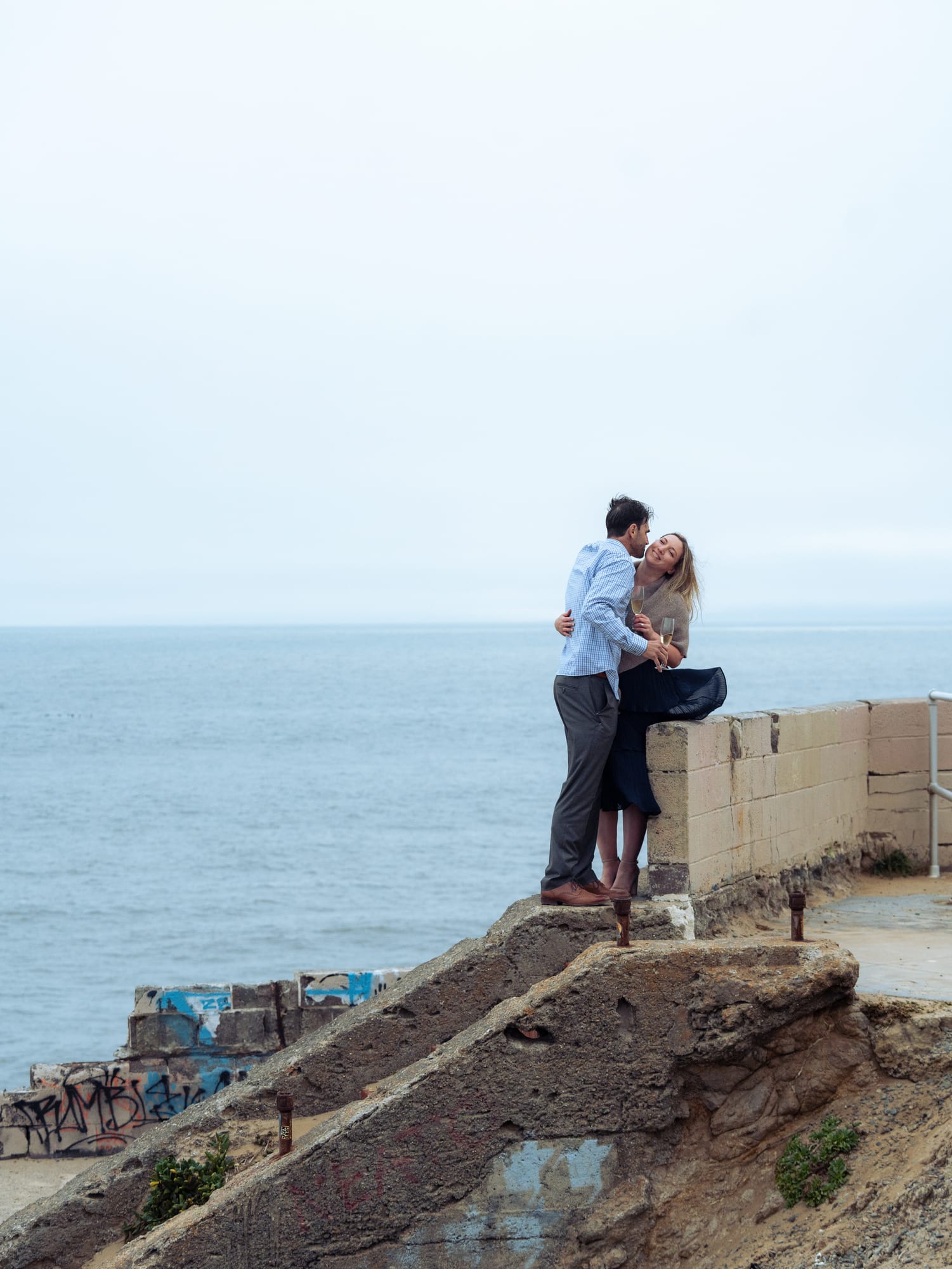 Couple standing together on the ruined seawall, arms wrapped around each other, the open ocean behind them