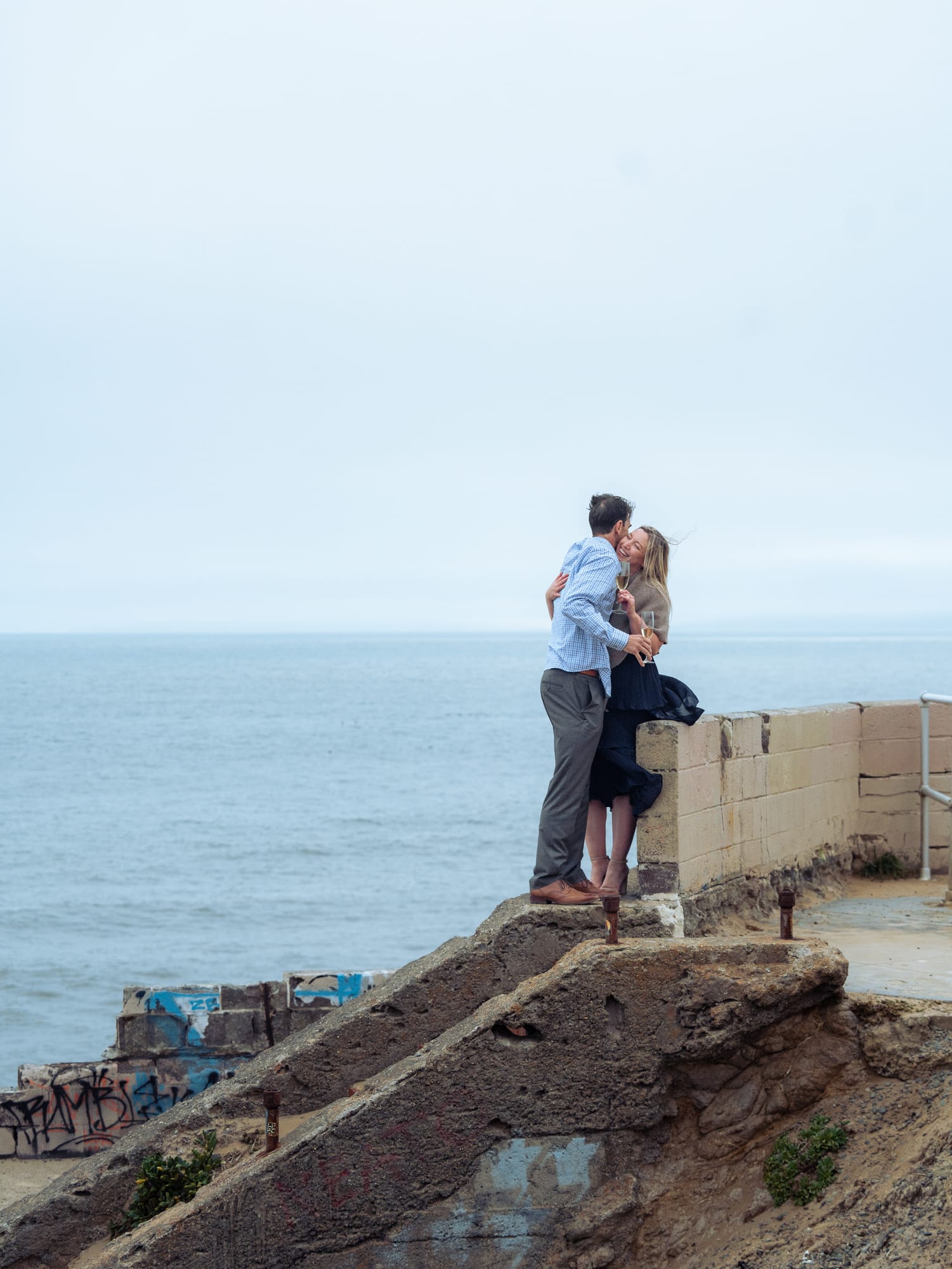 Couple kissing at the edge of the seawall, champagne glasses in hand, the graffitied ruins below