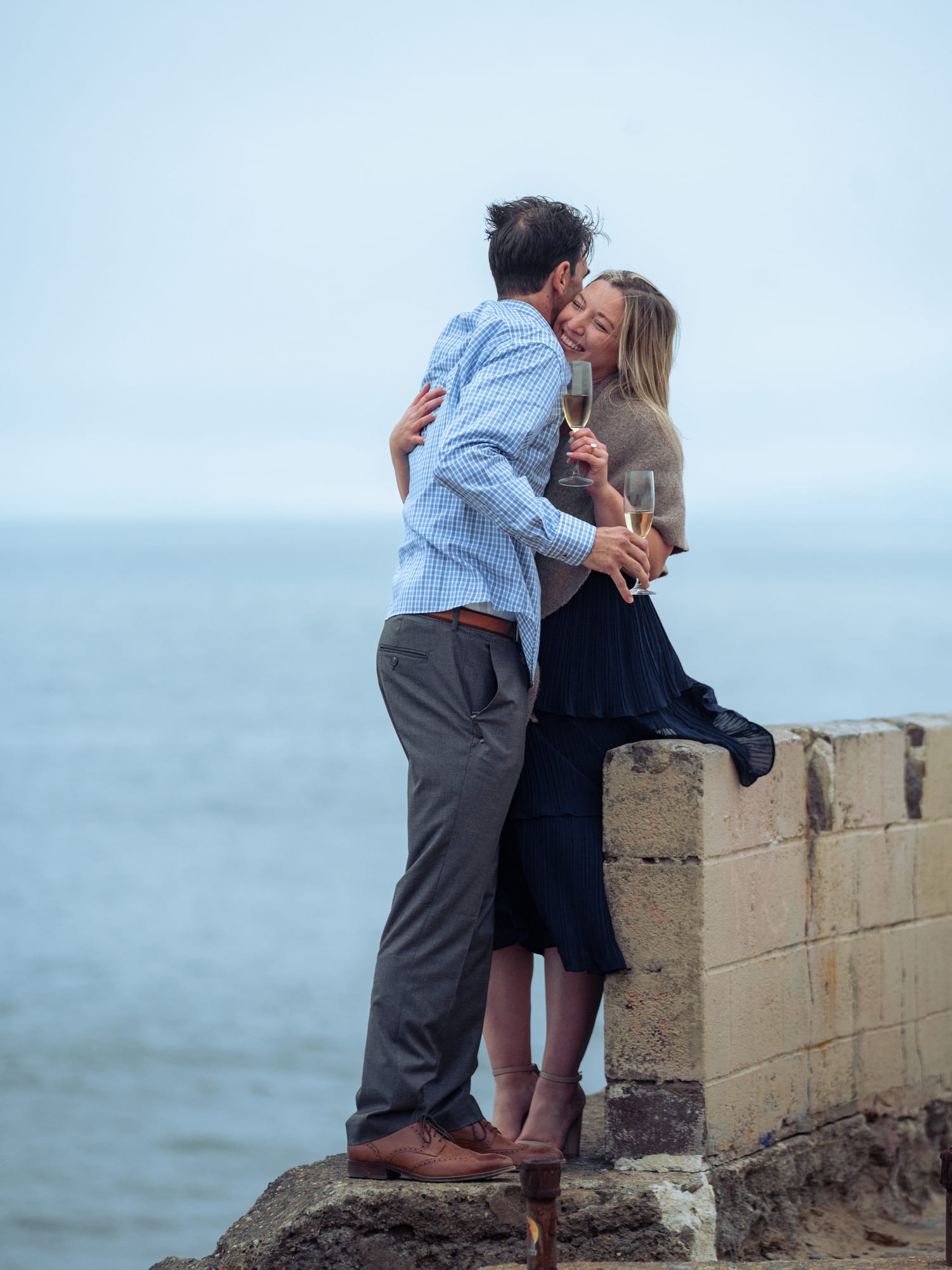 Couple laughing together at the edge of the seawall, holding champagne, the coastline curving away behind them