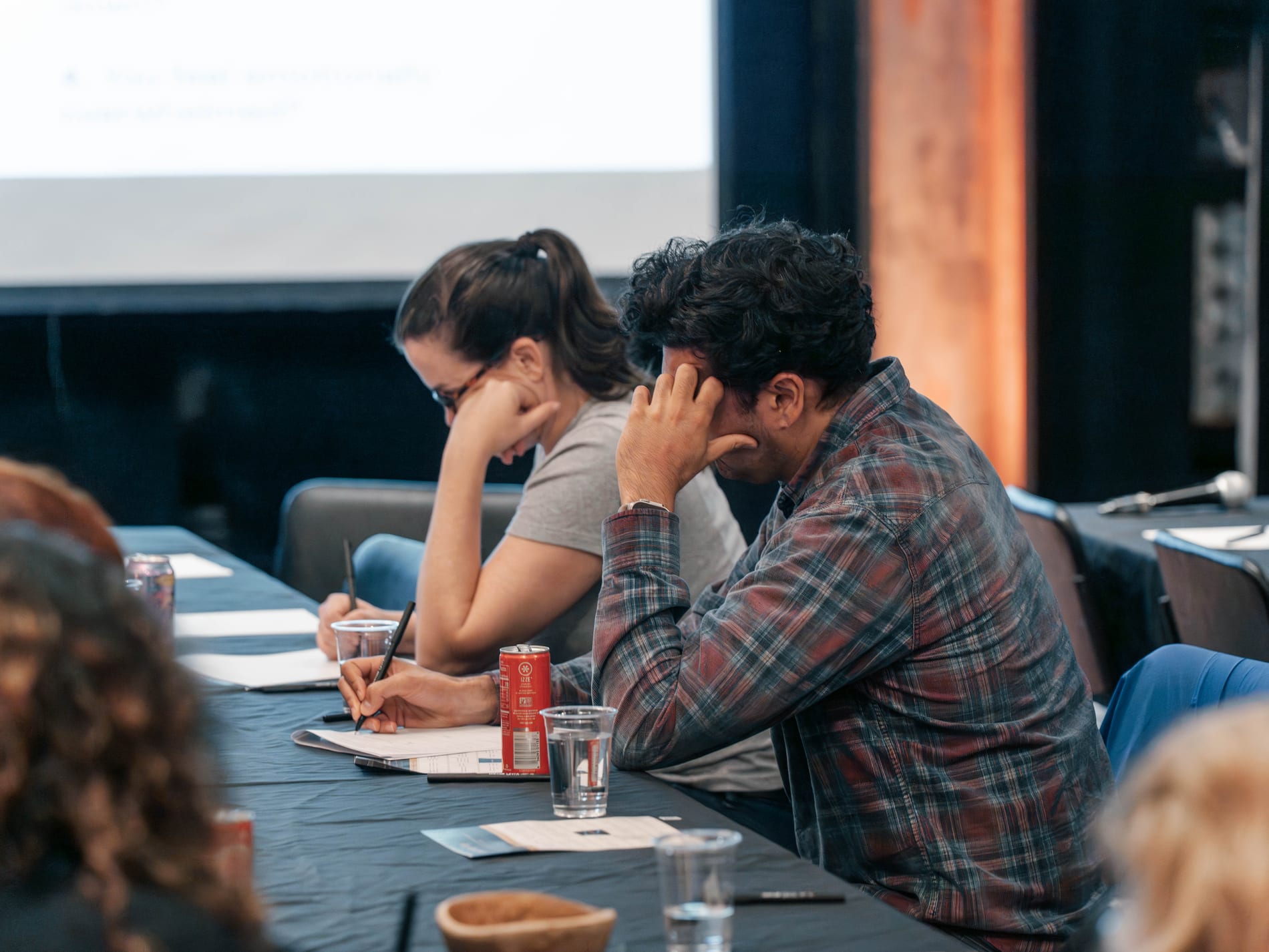 A workshop attendee presses his fingers to his temple while the guest beside him takes notes at the conference table, a presentation screen glowing behind them