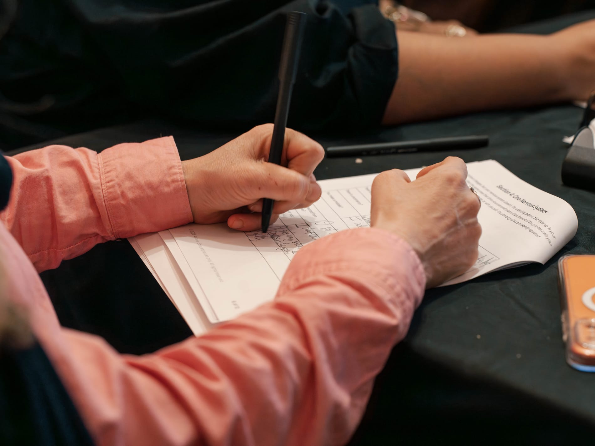 A guest in a pink sleeve fills out a workshop worksheet by hand, a pen and water glass resting on the table beside her