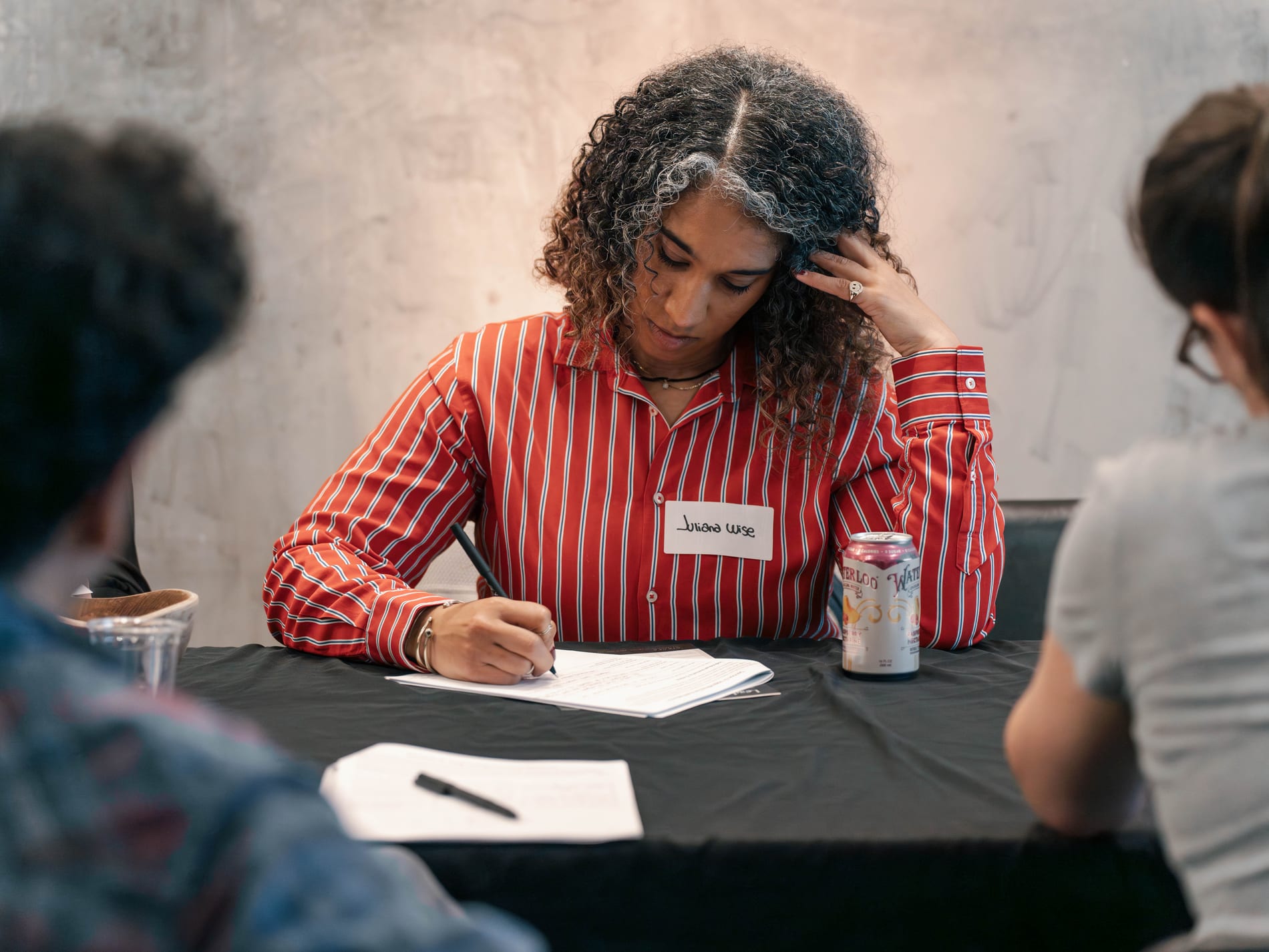 A guest in a red striped shirt and name tag jots notes on a worksheet at the table, a seltzer can and glass of water beside her