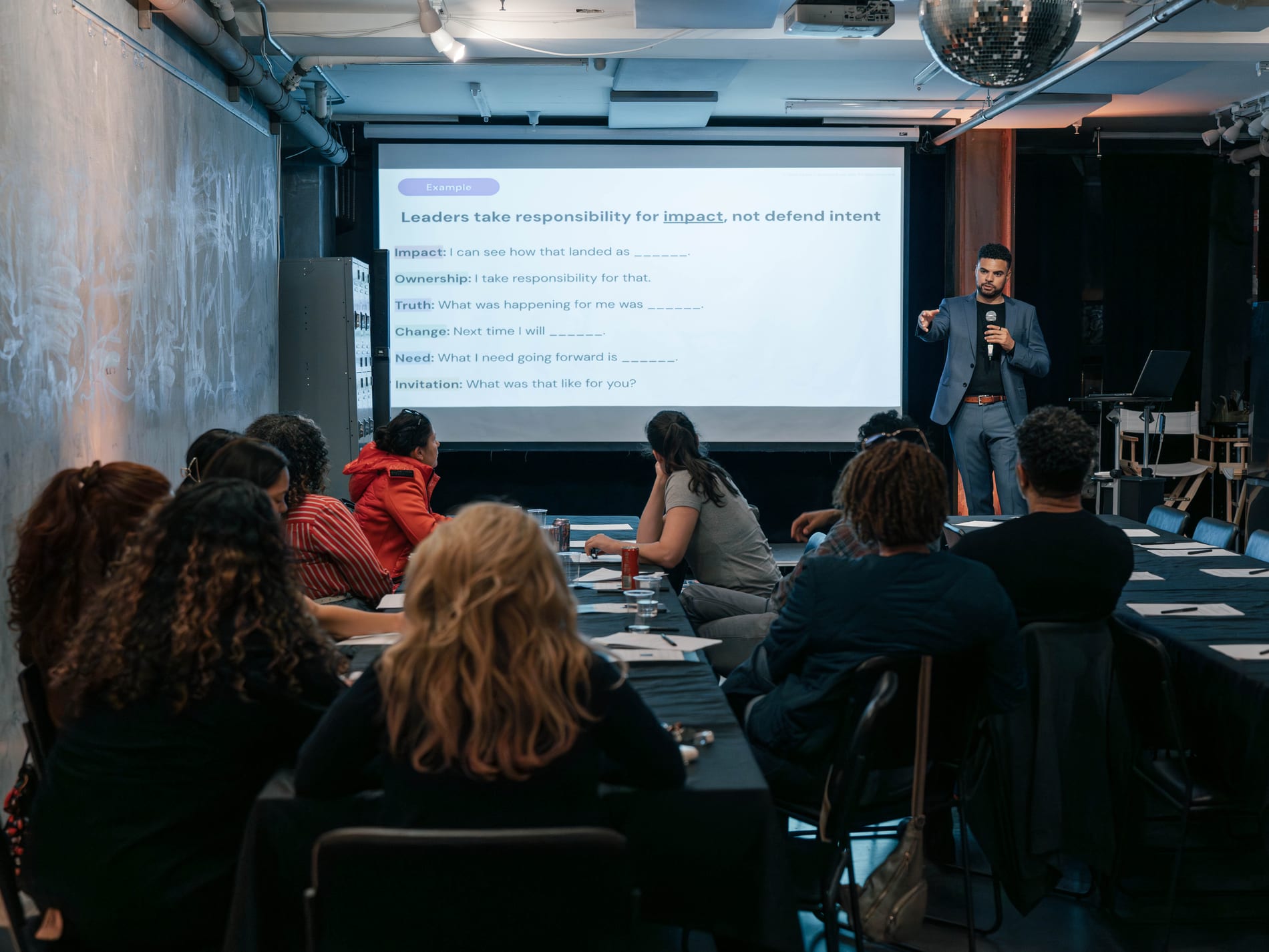 A speaker gestures with a microphone beside a screen reading Leaders take responsibility for impact, not defend intent, as guests take notes at the long tables below a disco ball