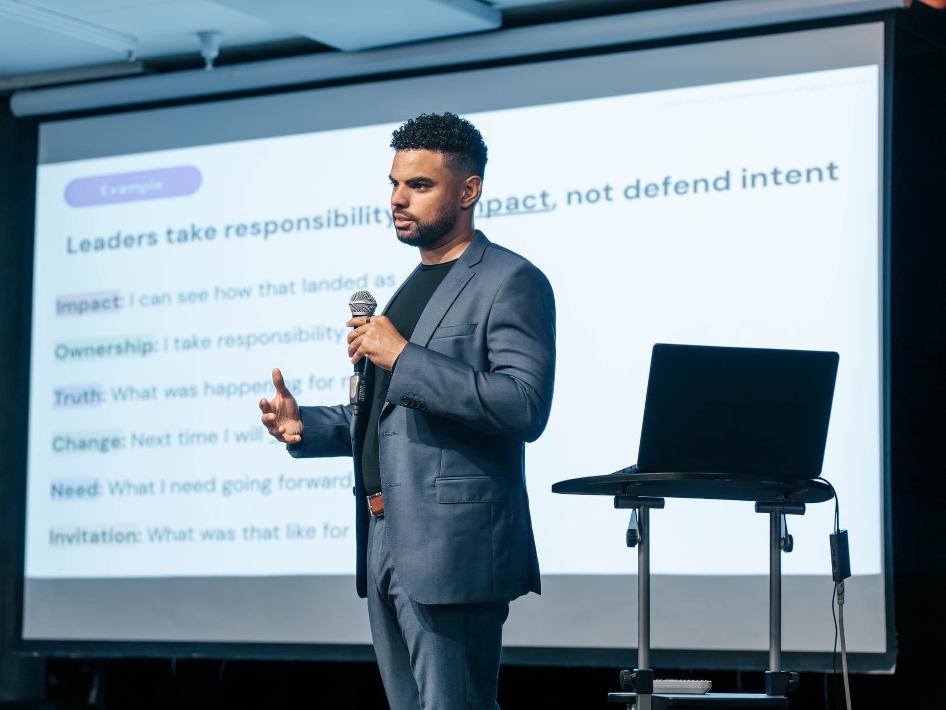 A speaker gestures mid-sentence with a microphone in hand, a laptop on a stand beside him and the slide Leaders take responsibility for impact, not defend intent lit up on the screen behind
