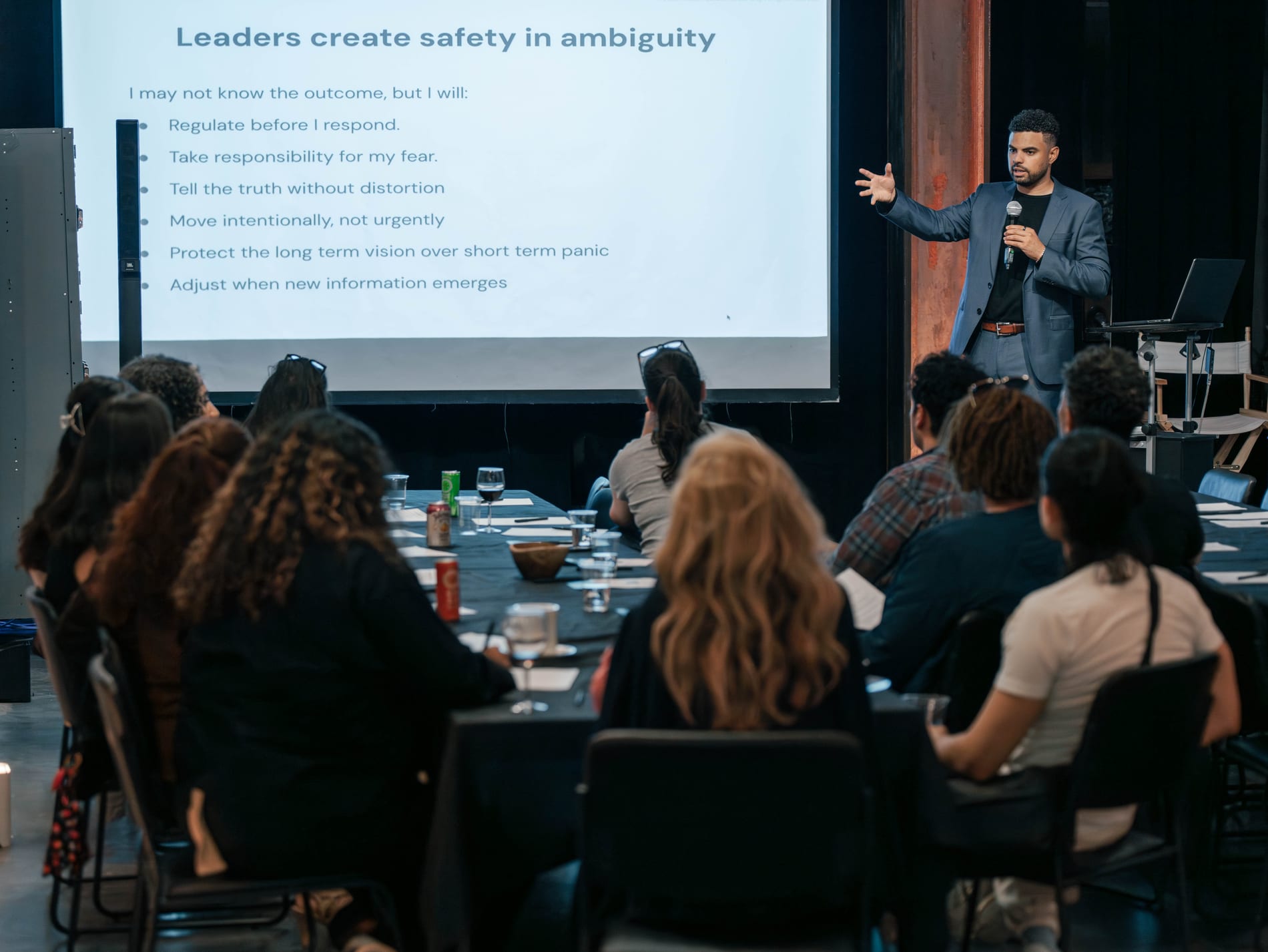A speaker gestures with an open hand beside a screen reading Leaders create safety in ambiguity, guests seated at the table with wine and water glasses in front of him