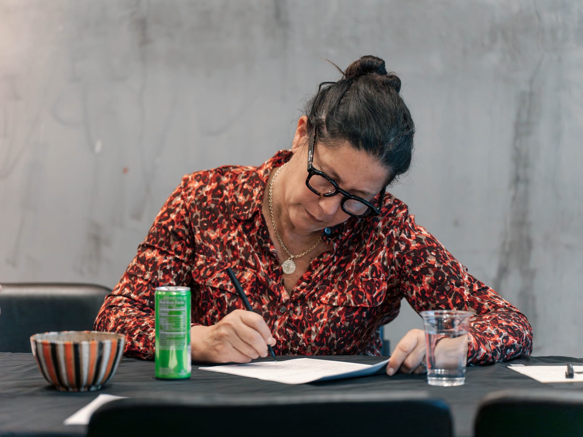 A guest in a leopard print blouse writes on a worksheet at the table, a green seltzer can and striped ceramic bowl in front of her