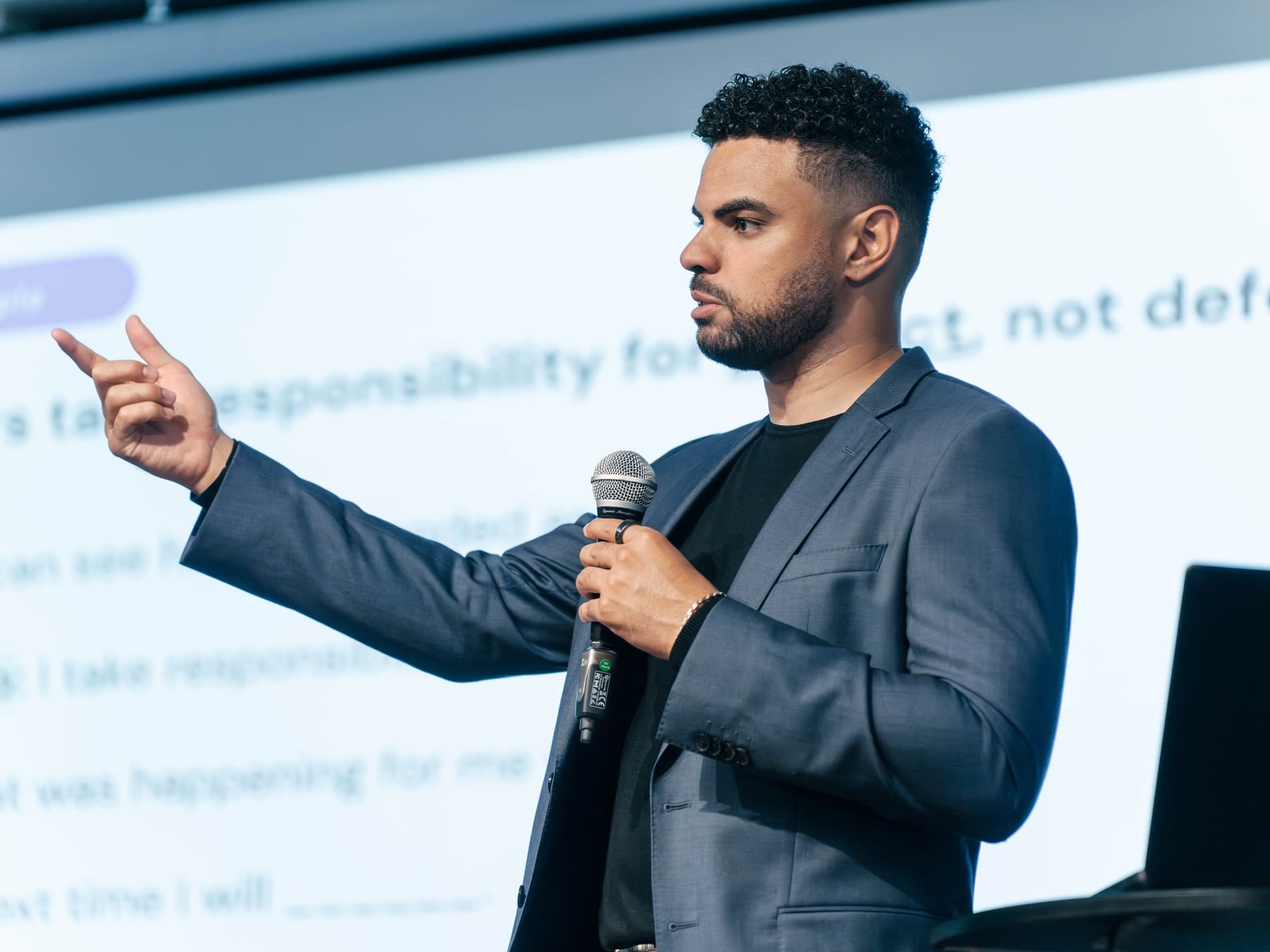 A speaker points out across the room with a microphone in hand, the slide filling the wall behind him reading Leaders take responsibility for impact, not defend intent