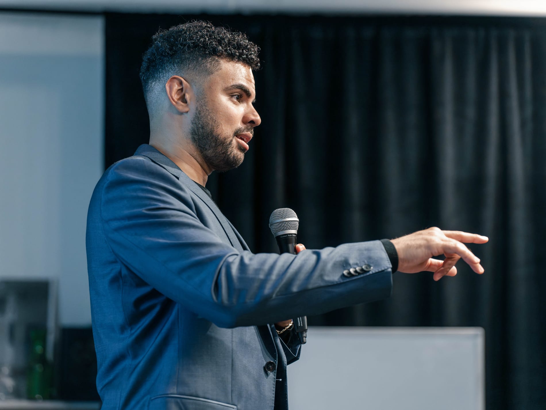 A speaker in a blue blazer reaches out with an open hand while holding a microphone, a dark stage curtain behind him