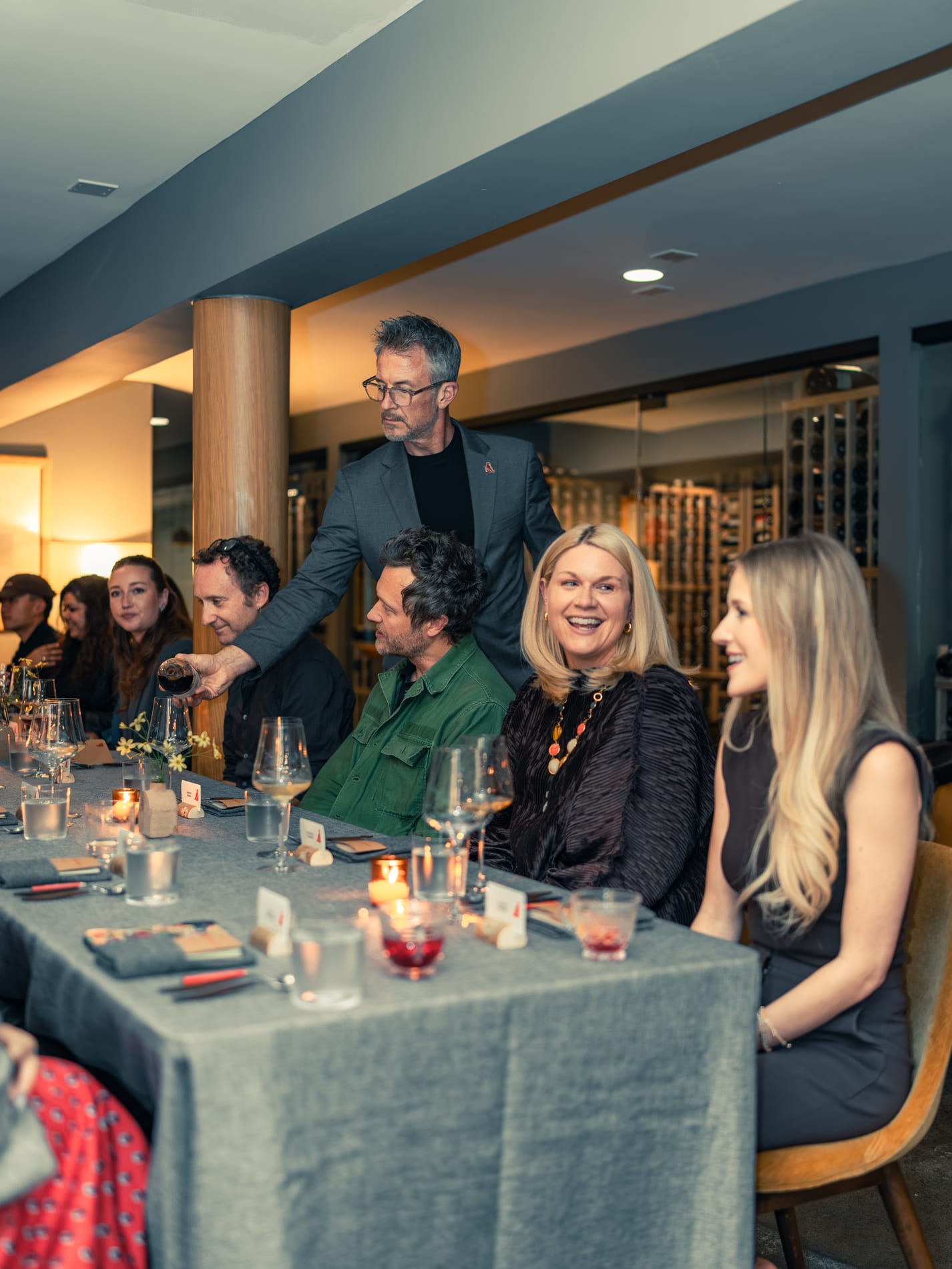 A host in a grey blazer pours wine over the shoulder of seated guests while two women laugh at a long dinner table, a lit wine cellar glowing behind them