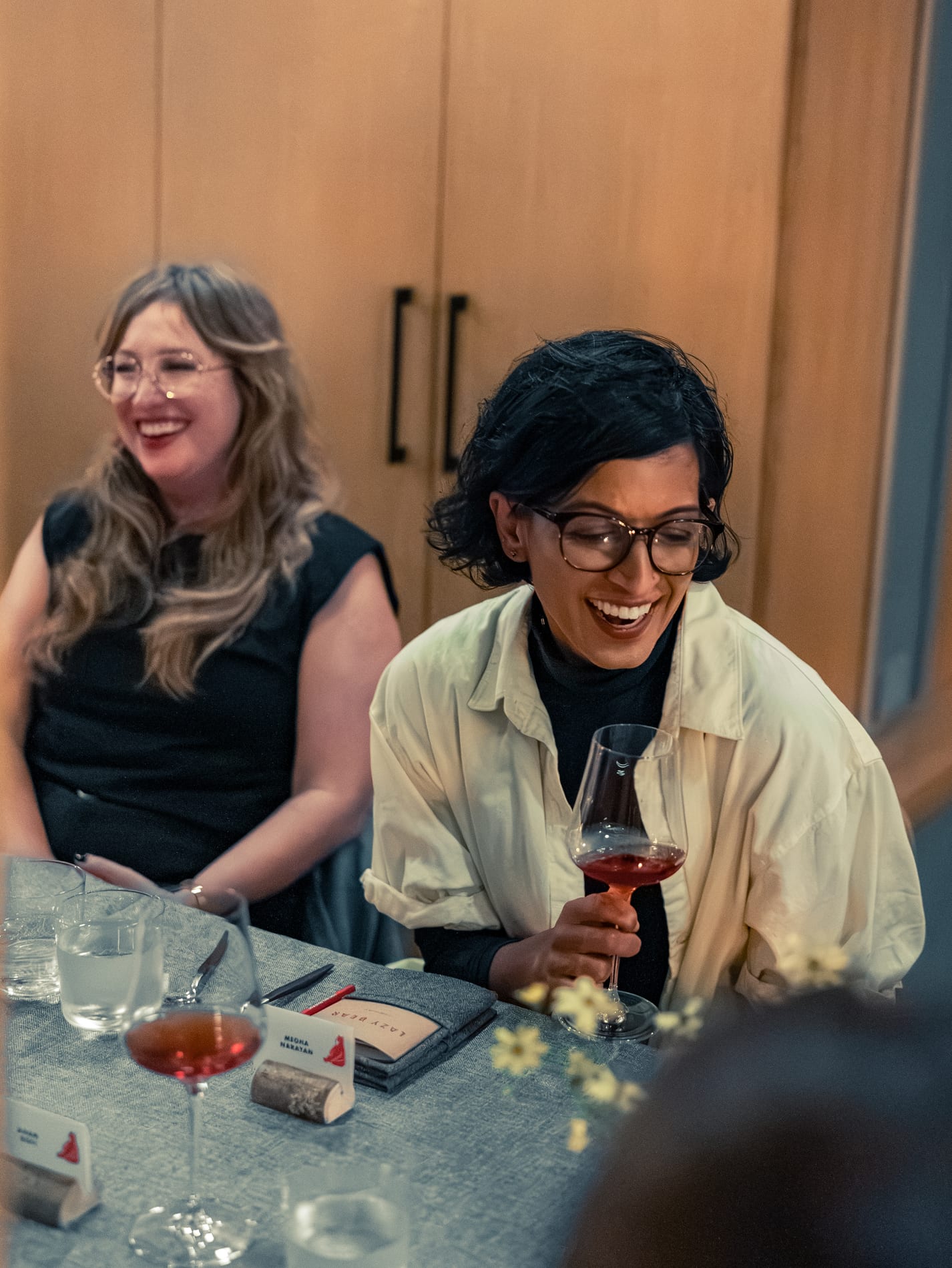 Two guests laugh at the table, one holding a glass of red wine, a place setting card reading Lazy Bear in the foreground