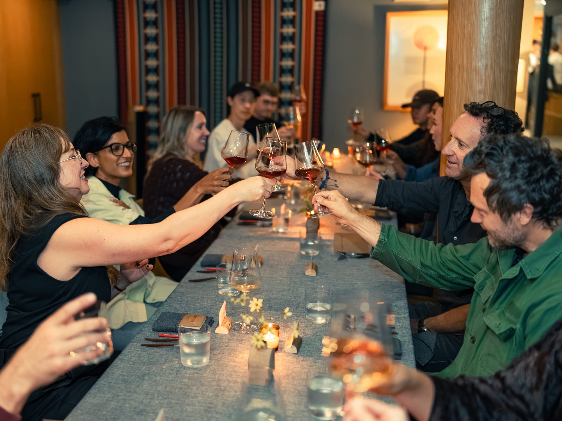 Guests lean across a long table to clink wine glasses together, candlelight and a striped woven wall hanging behind them