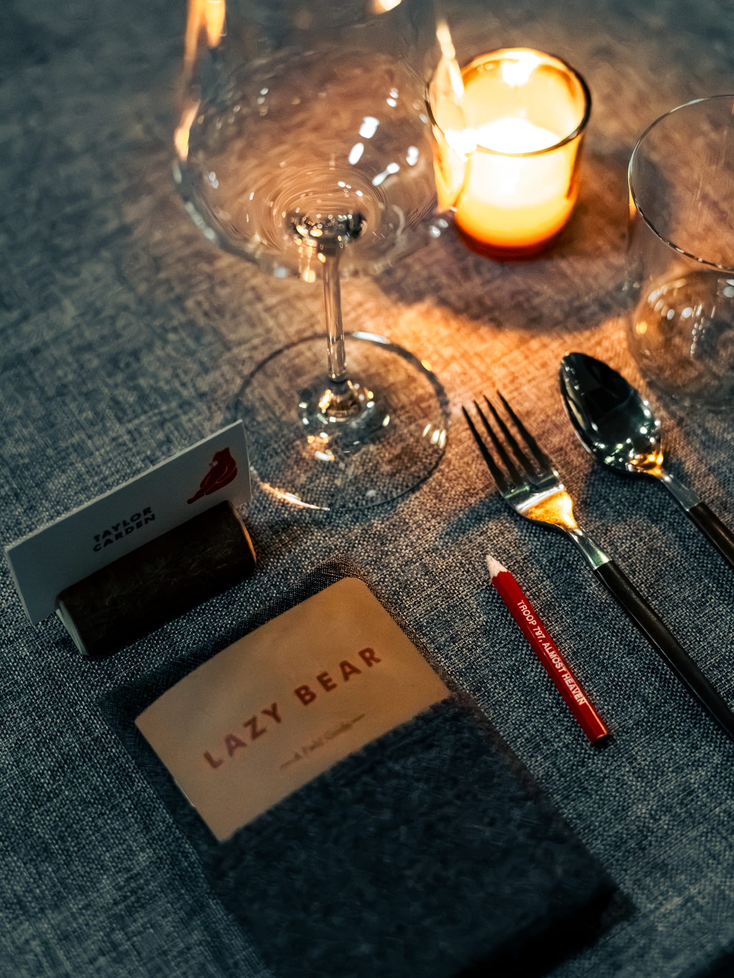 A wine glass, a lit candle and a place setting card reading Lazy Bear arranged on the dinner table