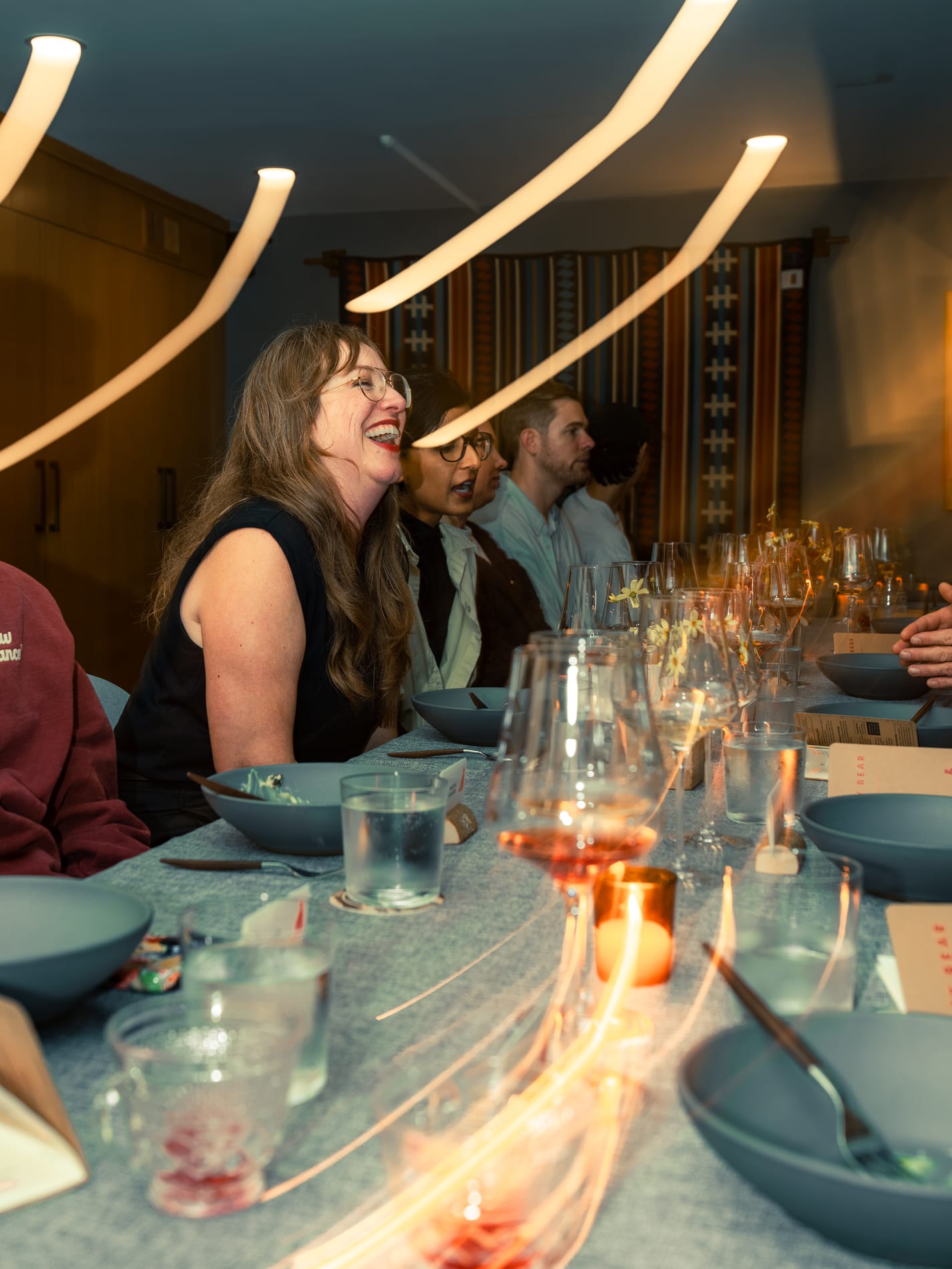 A guest laughs at the head of a long dinner table set with bowls and wine glasses, sculptural pendant lights glowing overhead