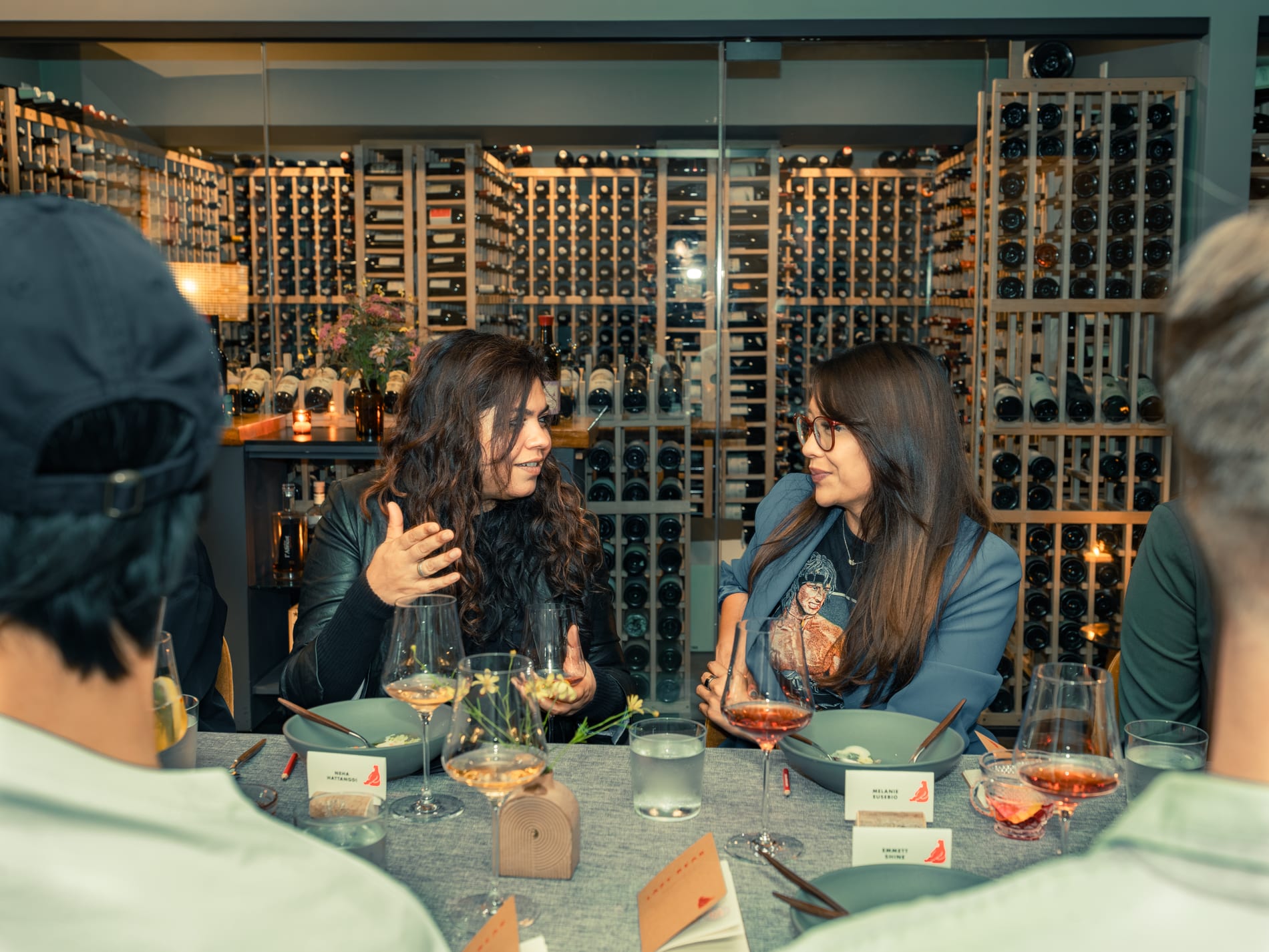 Two guests talk over dinner in a wine cellar, seated at a table set with wine glasses and small flowers among floor to ceiling wine racks