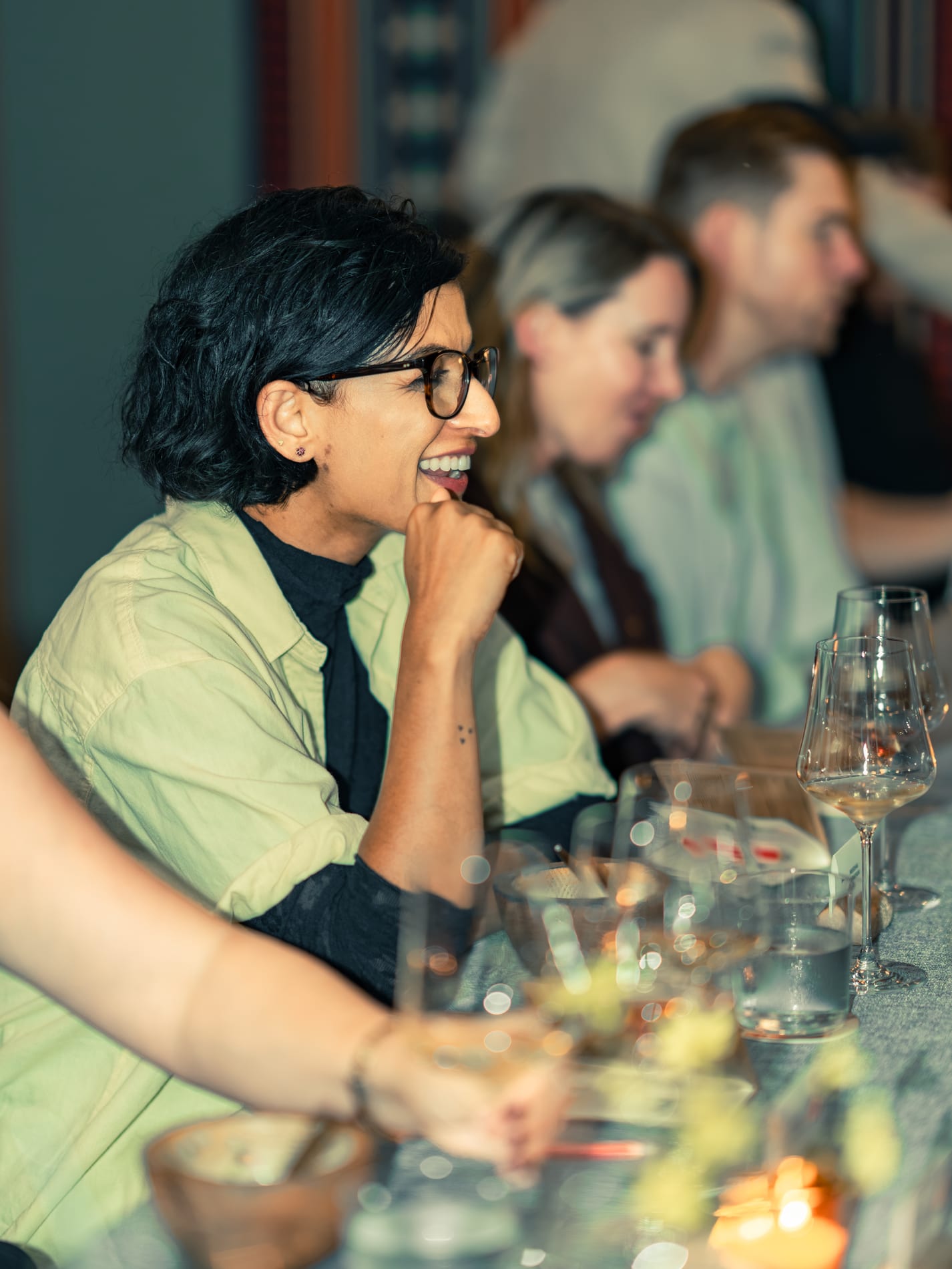 A guest in glasses smiles in profile with her chin resting on her hand, wine glasses blurred on the table before her