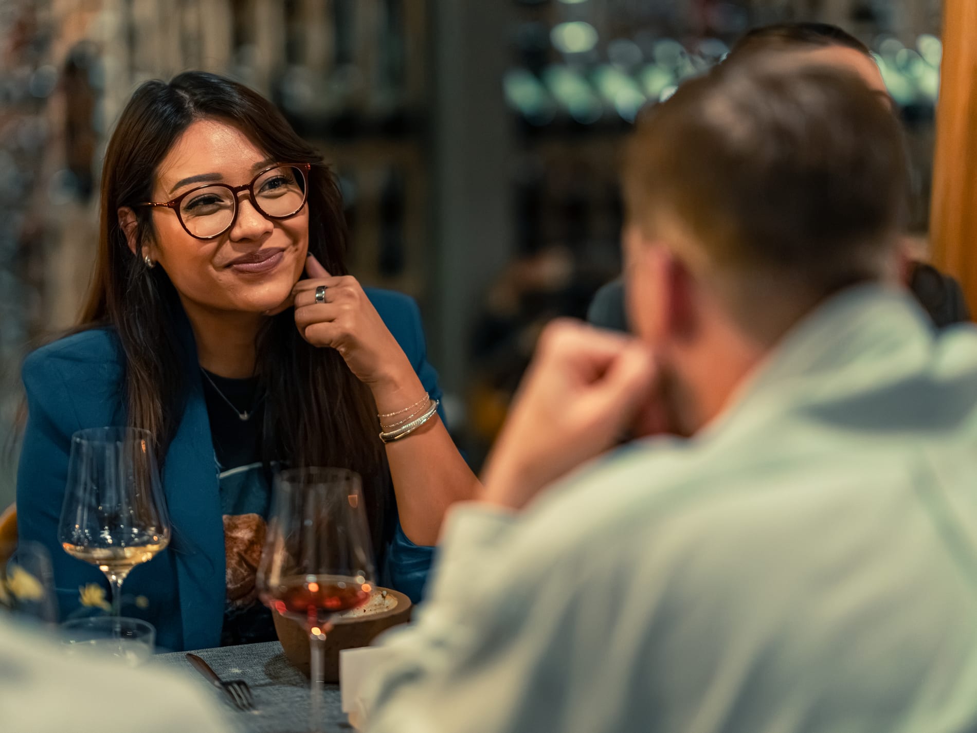 A guest in glasses smiles across the table at another guest, lit wine cellar shelves glowing behind her