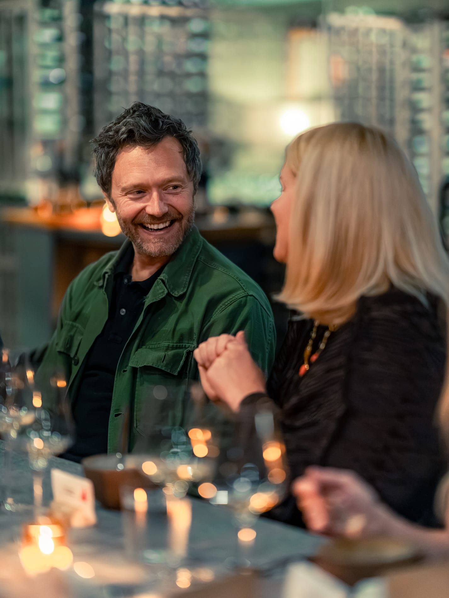 A man and woman laugh while clasping hands over the table, a lit city skyline visible through the window behind them