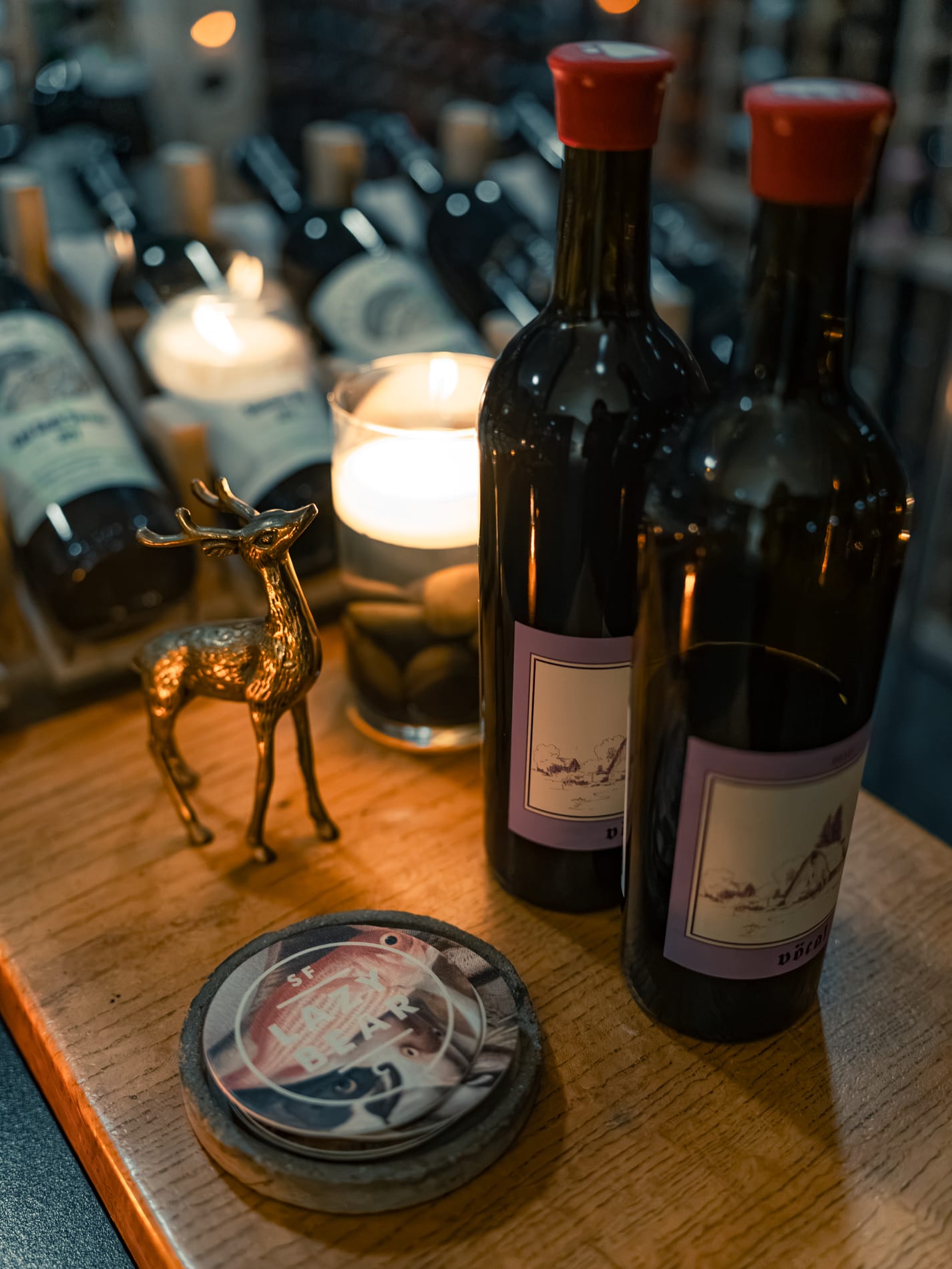 Two hand-labelled wine bottles stand beside a lit candle and a small bronze deer figurine, a coaster reading SF Lazy Bear in front