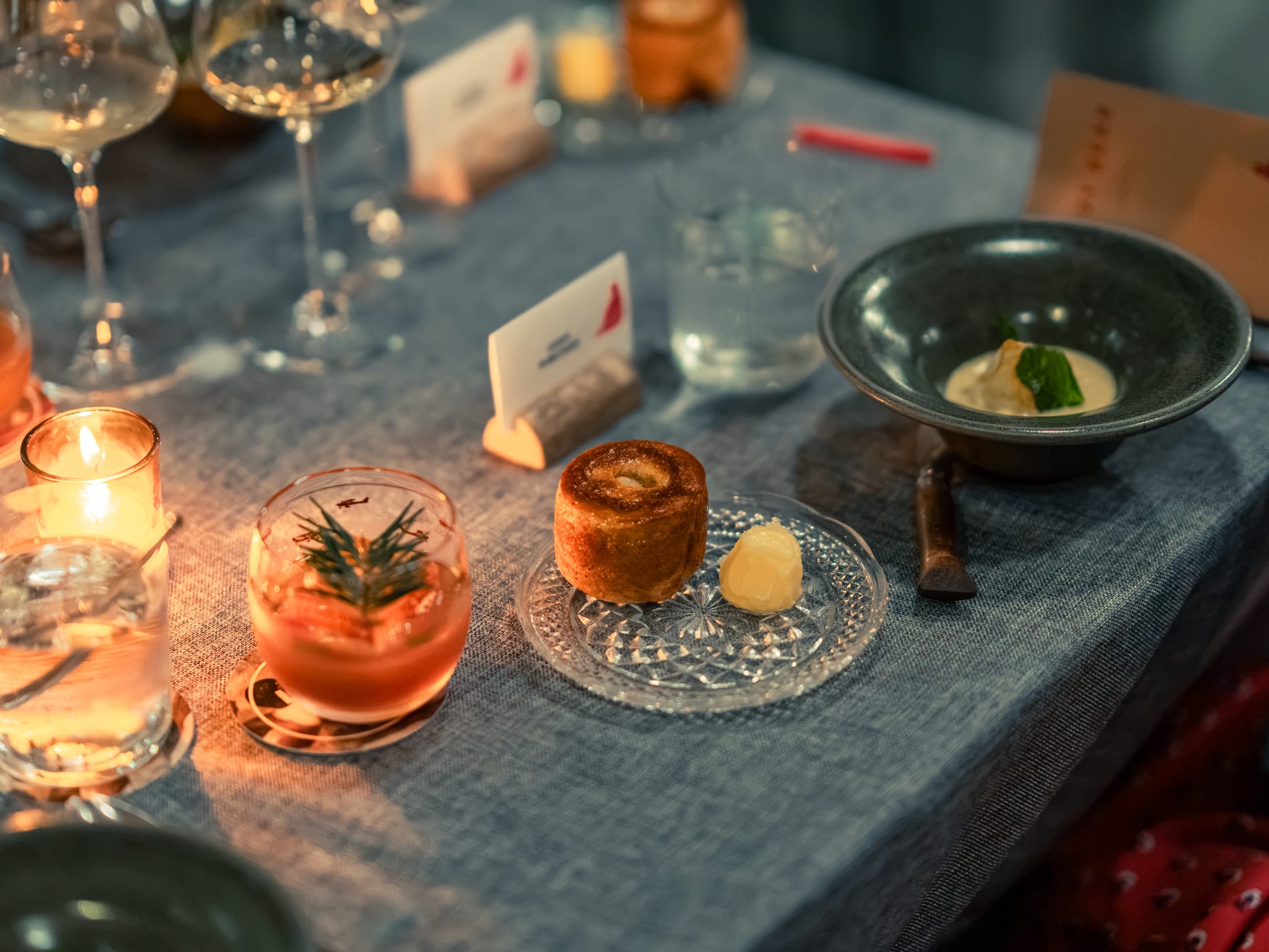 A savoury pastry with butter, a rosemary-garnished cocktail and a bowl of soup arranged on the candlelit table