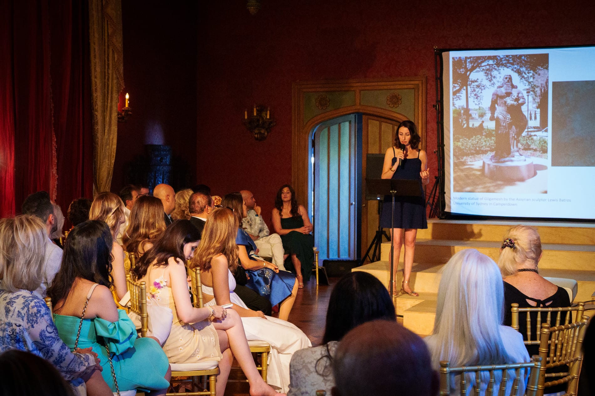 A speaker in a navy dress addresses a seated audience in a red walled mansion ballroom beside a projection screen showing a statue of Gilgamesh