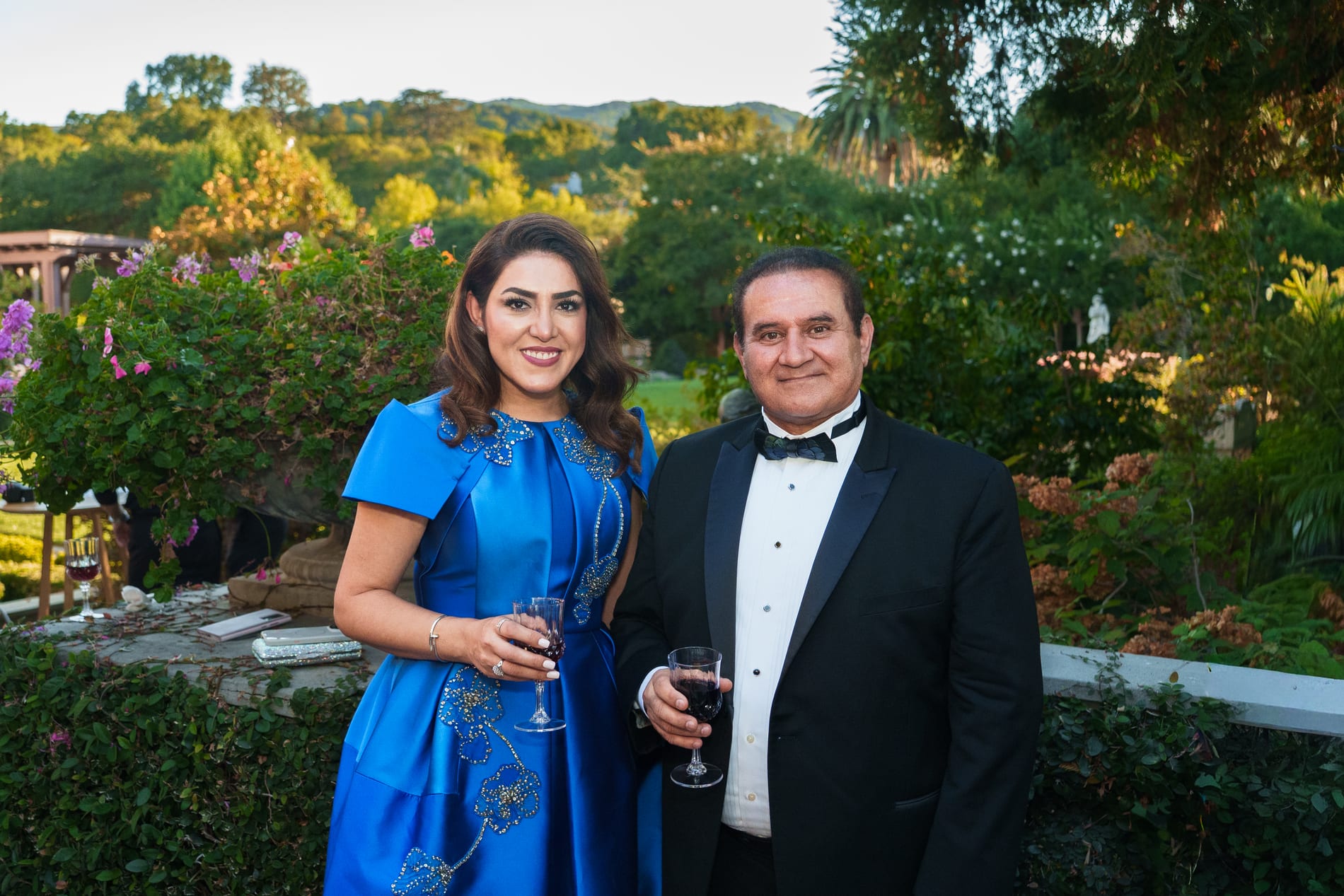 A couple in a beaded blue gown and black tuxedo hold glasses of red wine on a garden terrace overlooking wooded hills at dusk