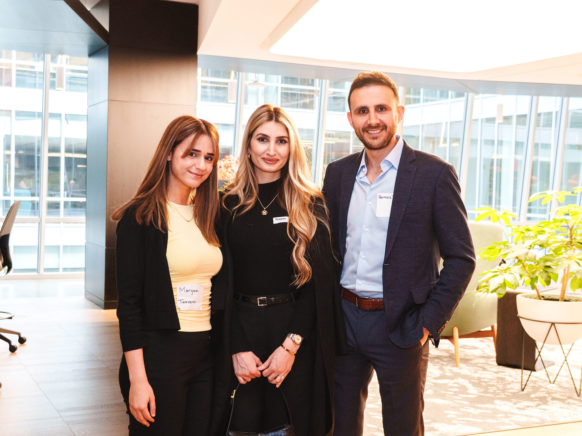 Three colleagues wearing name badges pose together in an office lobby with floor to ceiling windows behind them