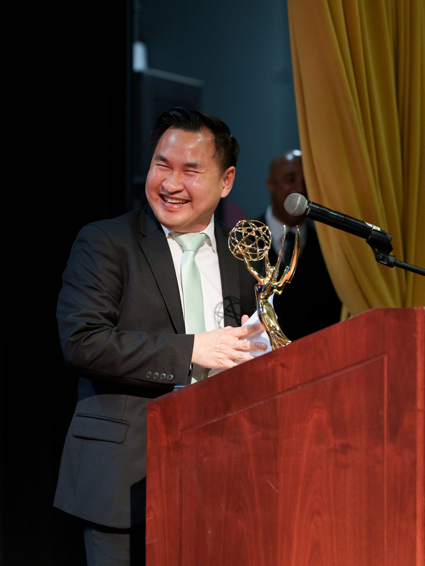 An award recipient in a suit and mint tie laughs while cradling his Emmy statuette at the podium, a gold curtain behind him