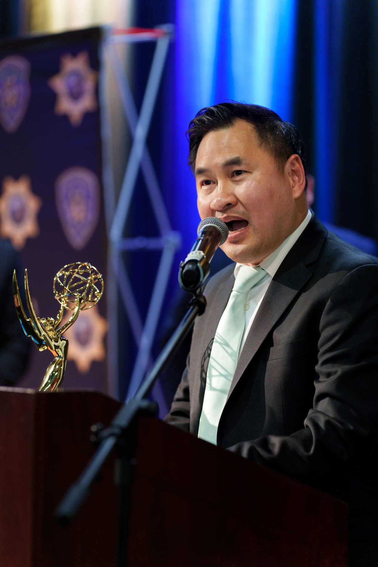 An award recipient speaks into the microphone at the podium, his Emmy statuette resting on the lectern in front of a lit blue curtain and a California State University Police badge backdrop