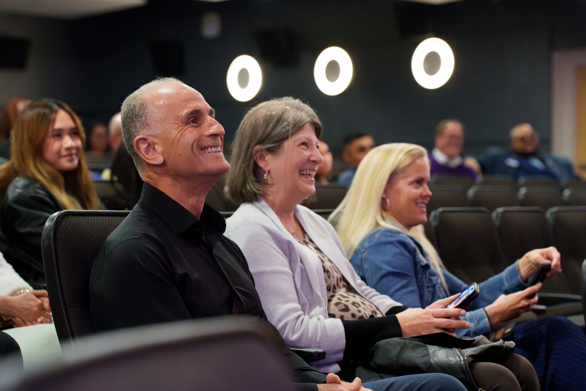 A row of seated guests laugh together during the ceremony, one holding up her phone to capture the moment
